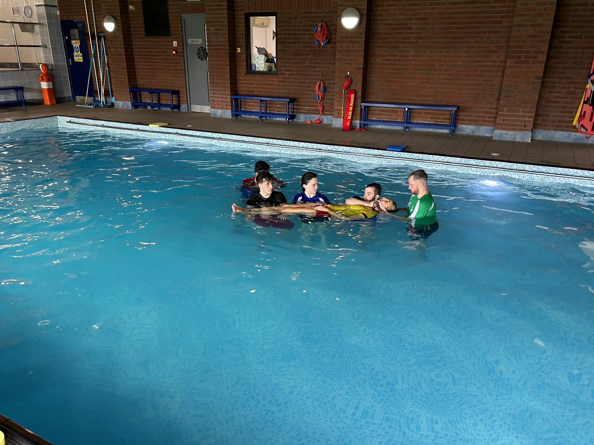 A group of people in a swimming pool, with one person being supported by others, during a water safety or rescue training session.