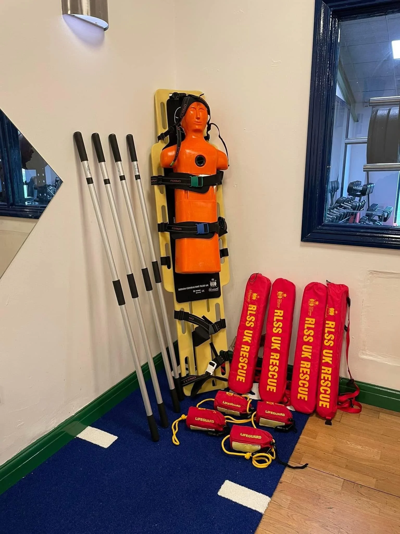 Rescue equipment including an orange mannequin torso with head and helmet, four yellow and black rescue stretchers labeled "RUSS UK RESCUE," four red rescue bags labeled "LIFEGUARD," and four walking poles, all arranged against a wall in a room.