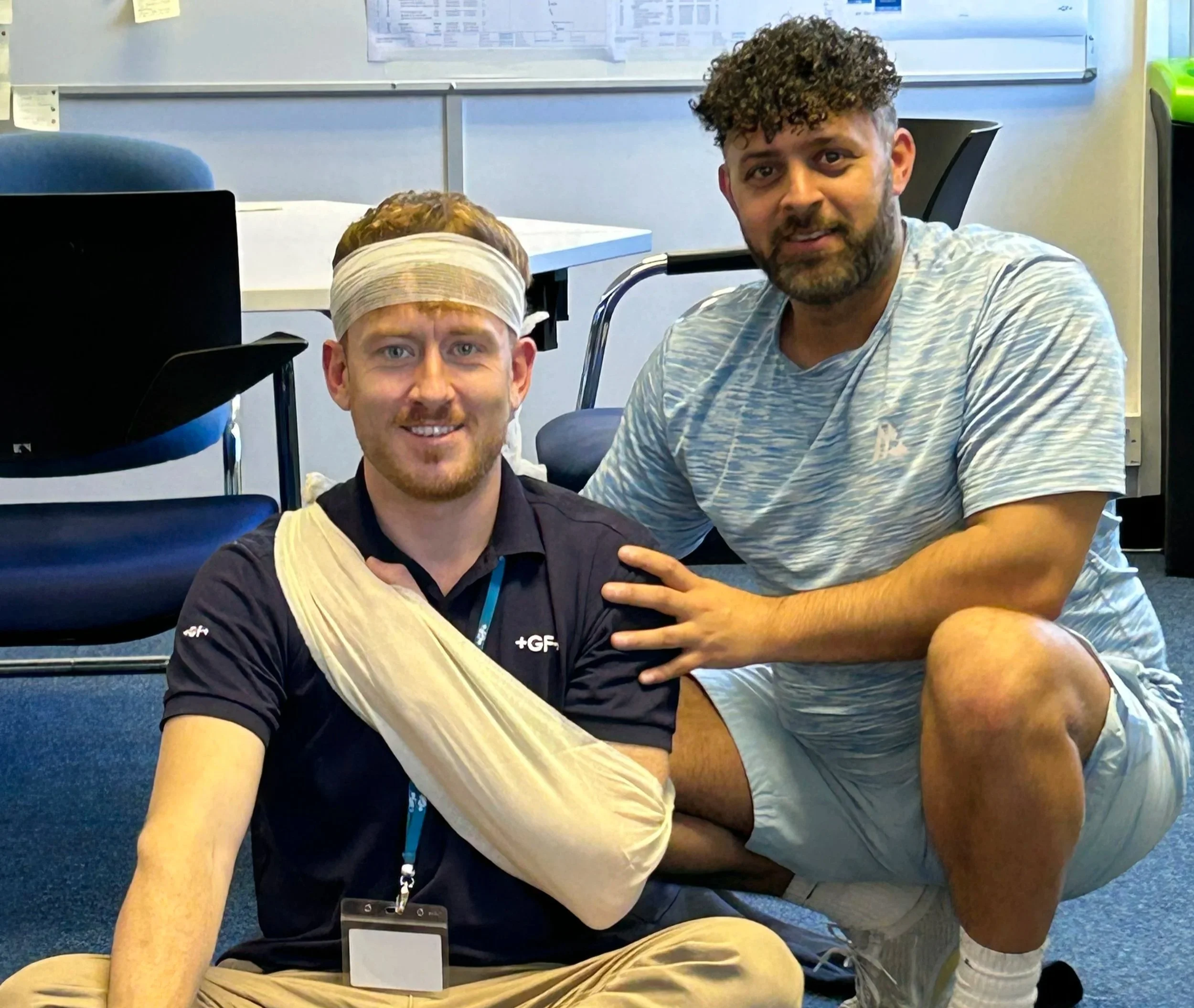 Two men sitting on the floor inside an office, one with a bandage and head injury, smiling at the camera, the other with his hand on the injured man's shoulder.