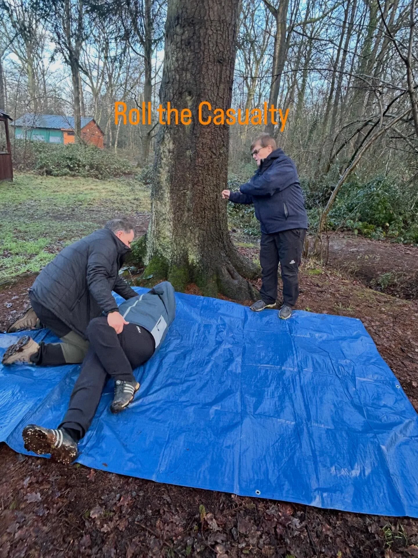 Two men are practicing a first aid scenario outdoors on a blue tarp in front of a large tree. One man is lying on the tarp, simulating a casualty, while the other is kneeling beside him. A third person is standing nearby, ready to assist. The setting