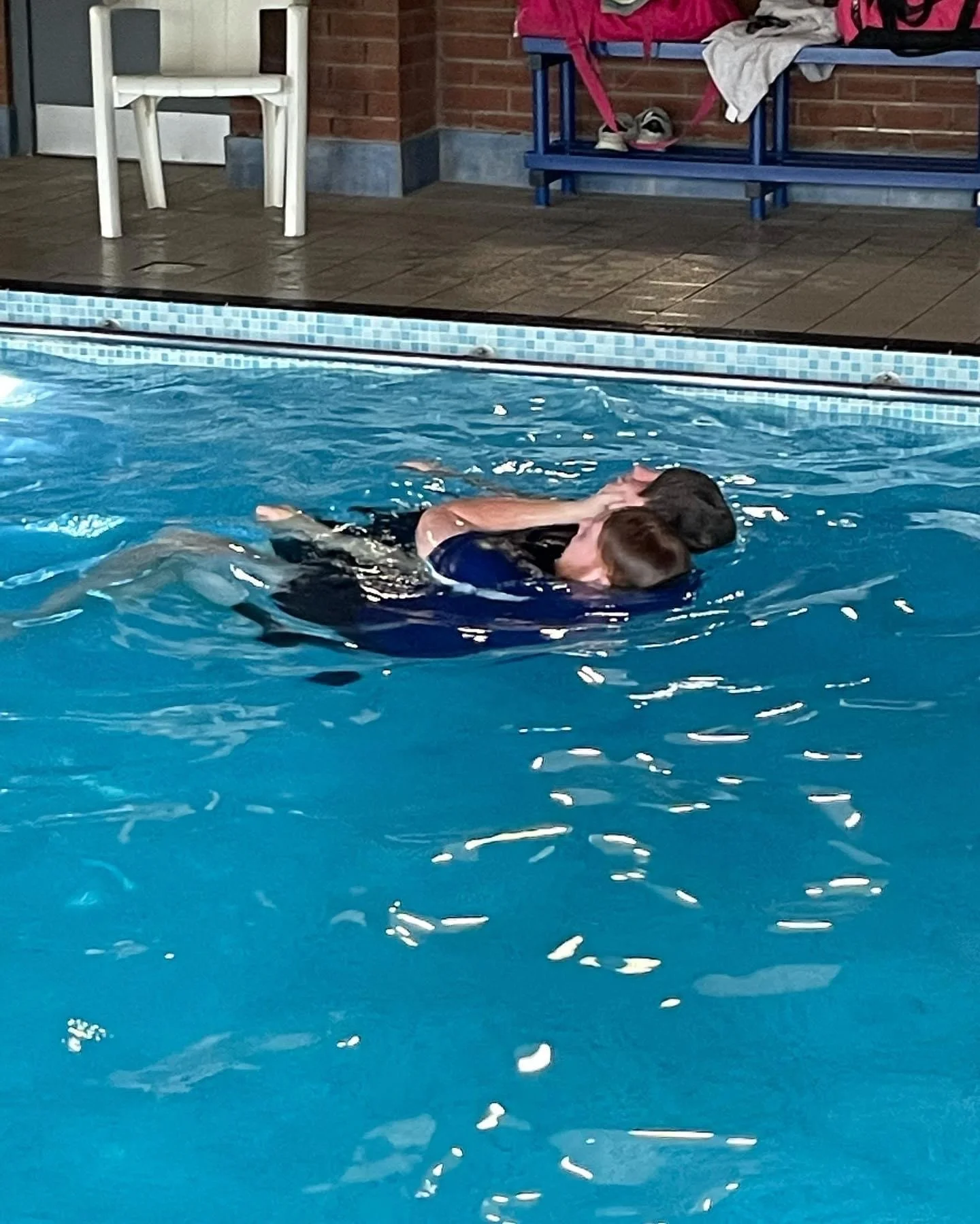 Two people swimming in a pool, one person holding another in an embrace. Poolside chairs and a bench with sports bags are visible in the background.