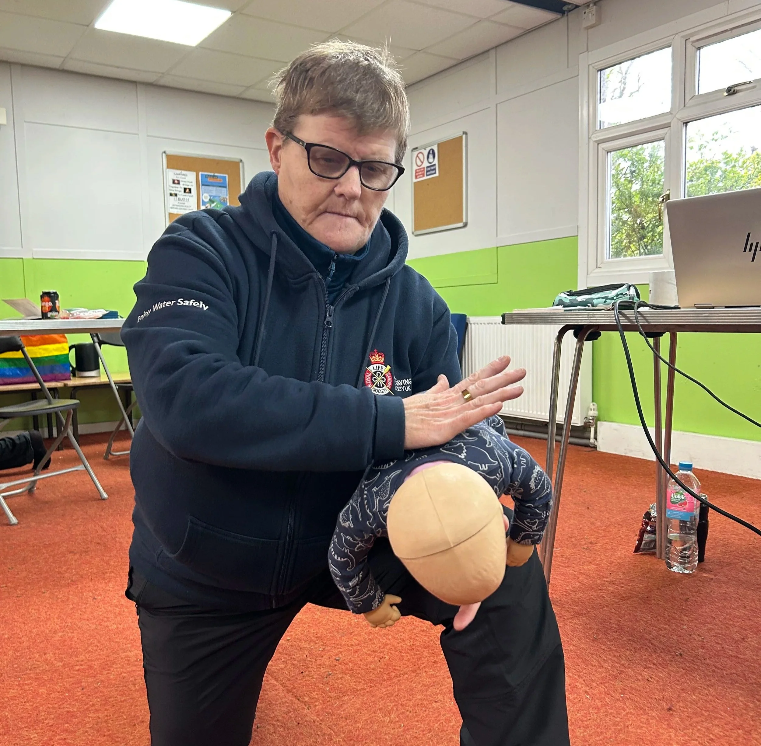 A person demonstrating CPR on a mannequin in a training room with green and white walls, windows, and training equipment.
