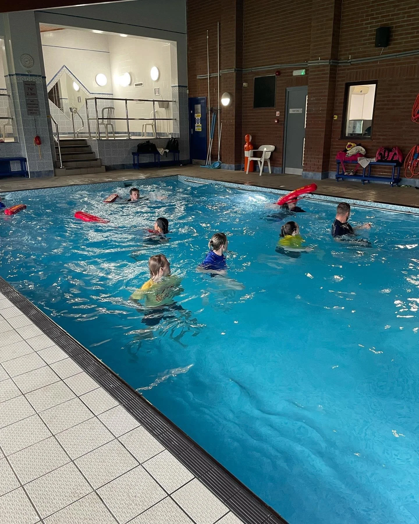 Indoor swimming pool with children receiving swimming lessons, some with floatation devices, in a well-lit room with tiled floors and walls, and safety equipment on the walls.