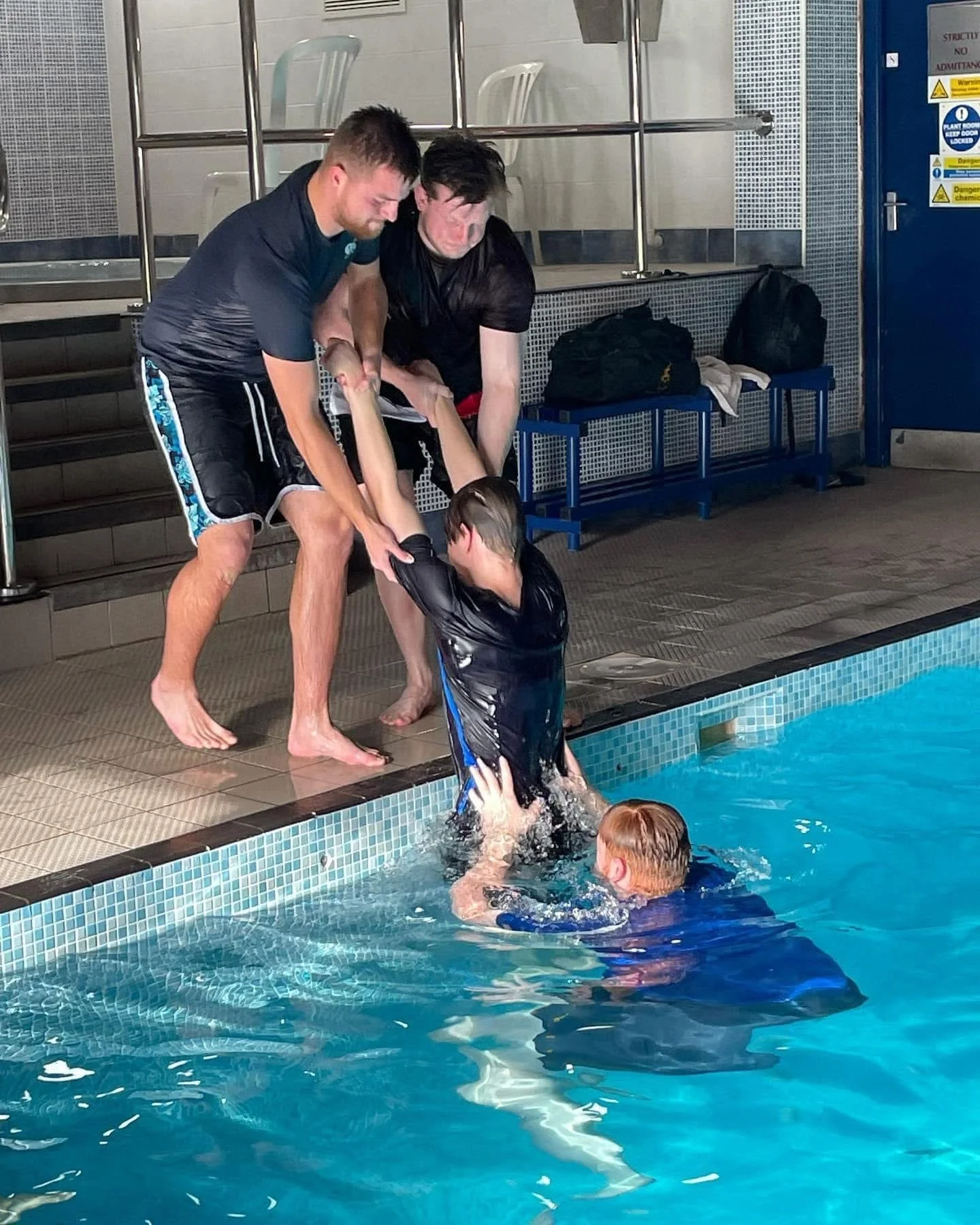 Two men assist a young girl in drowning, with a boy in the pool helping her, at an indoor swimming pool.