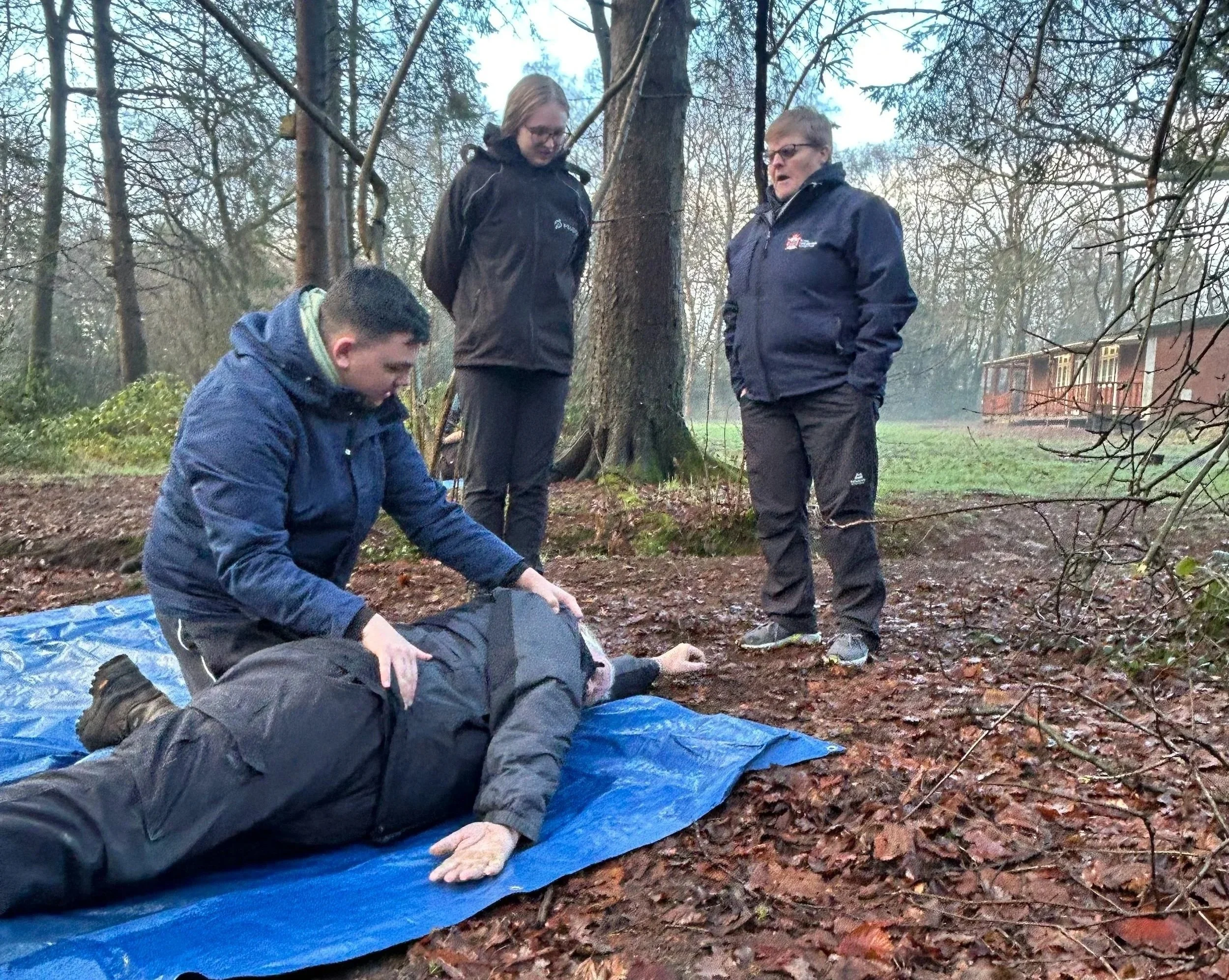 Three people practicing first aid on a mock casualty lying on a blue tarp in a wooded area.