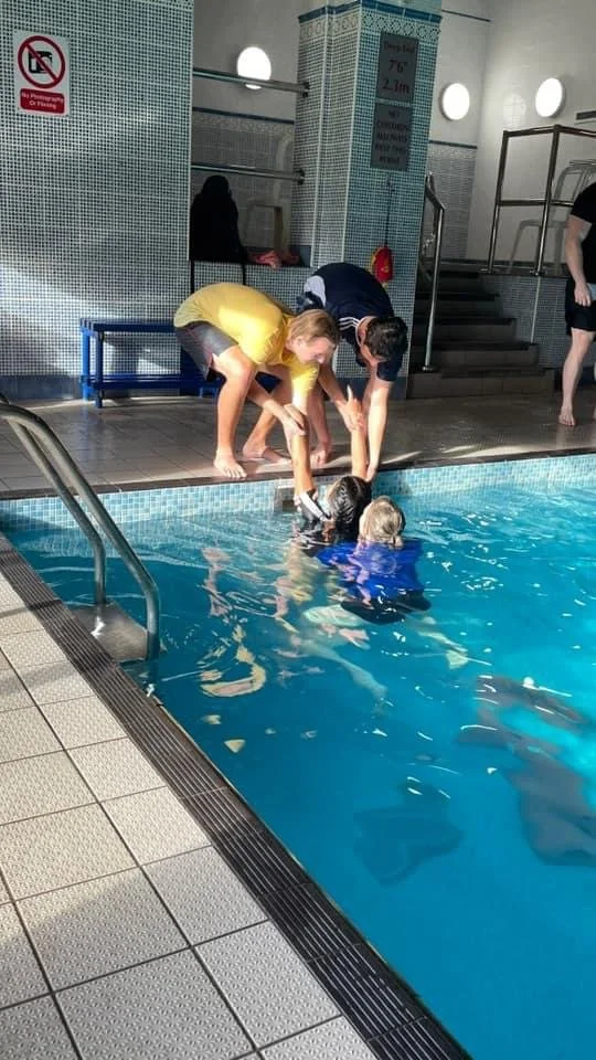 Two individuals are assisting two children in the swimming pool at an indoor facility. The helpers are reaching into the water to support the children, who are wearing blue swimsuits. The pool area has tiled walls and a staircase in the background.