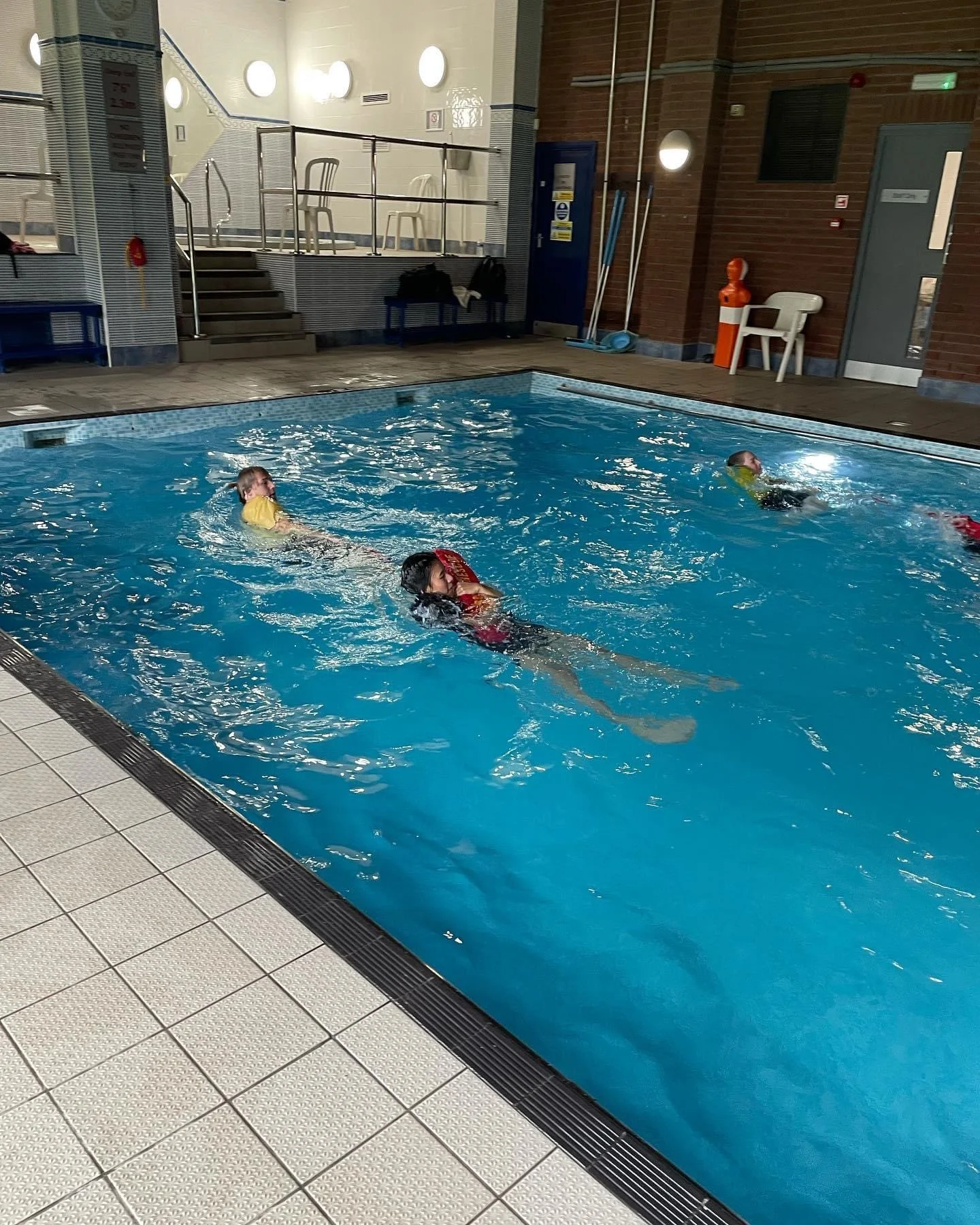 Indoor swimming pool with three children swimming, one near the wall, one in the middle, and one farther away, with a brick and tiled interior and poolside area.