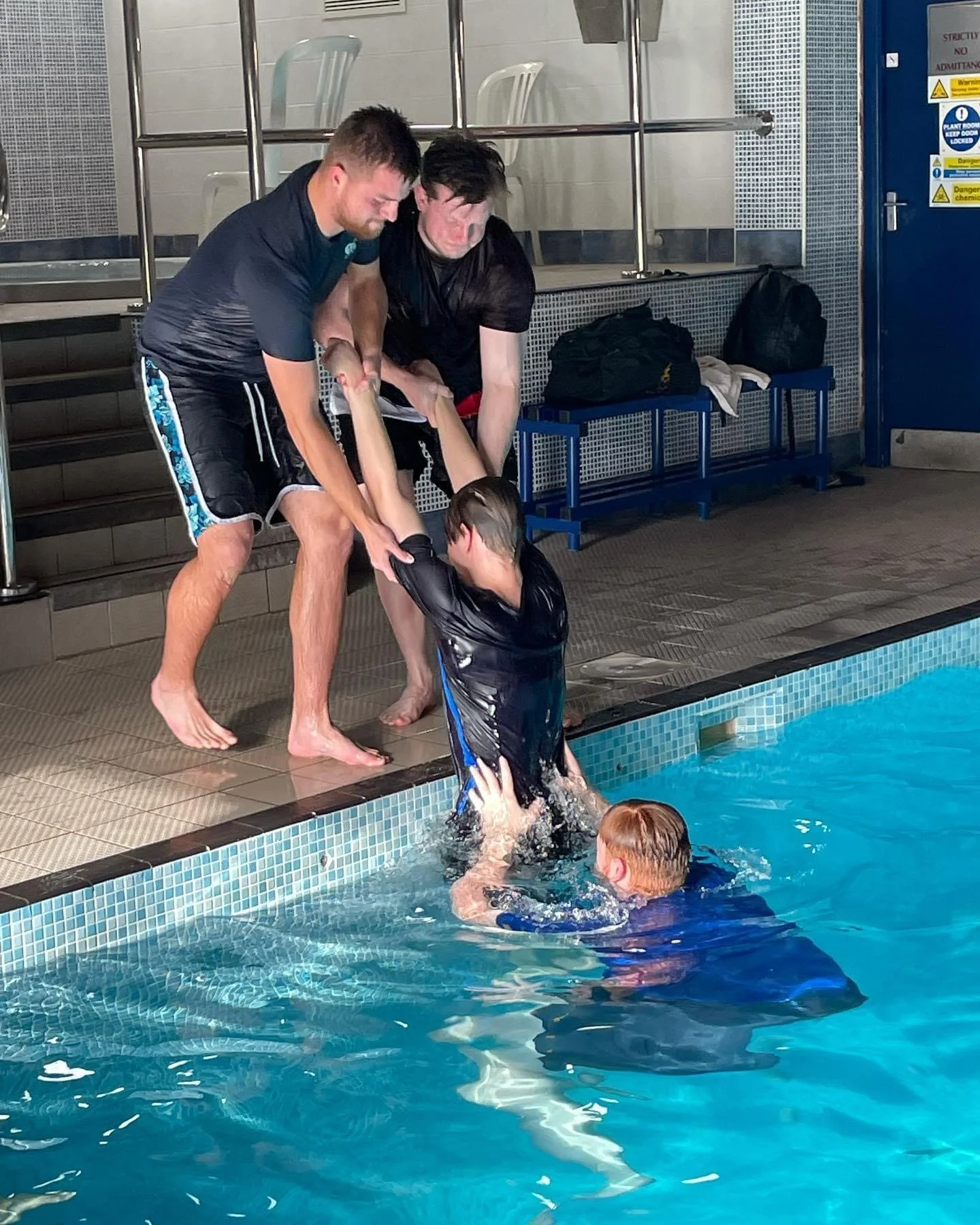 Two men assist a young boy in entering a swimming pool by holding his arms while another child supports him from below in the water.