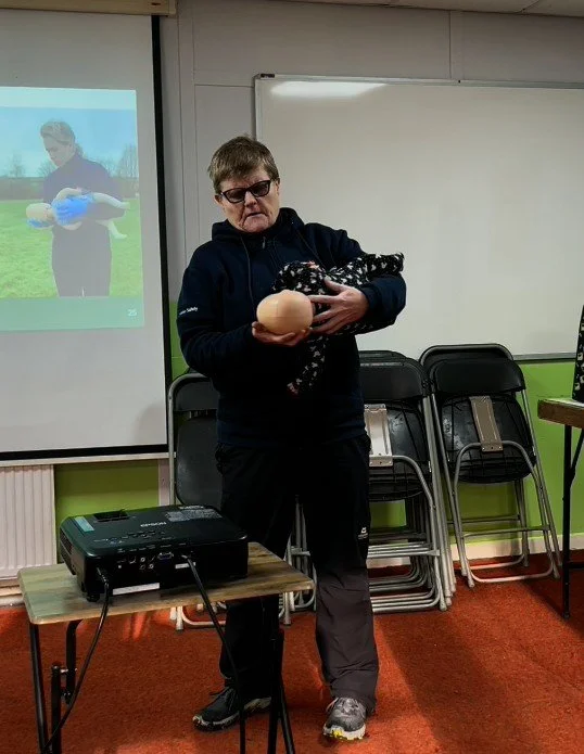 Woman wearing sunglasses holding a baby doll and an egg in a classroom with chairs and a projector