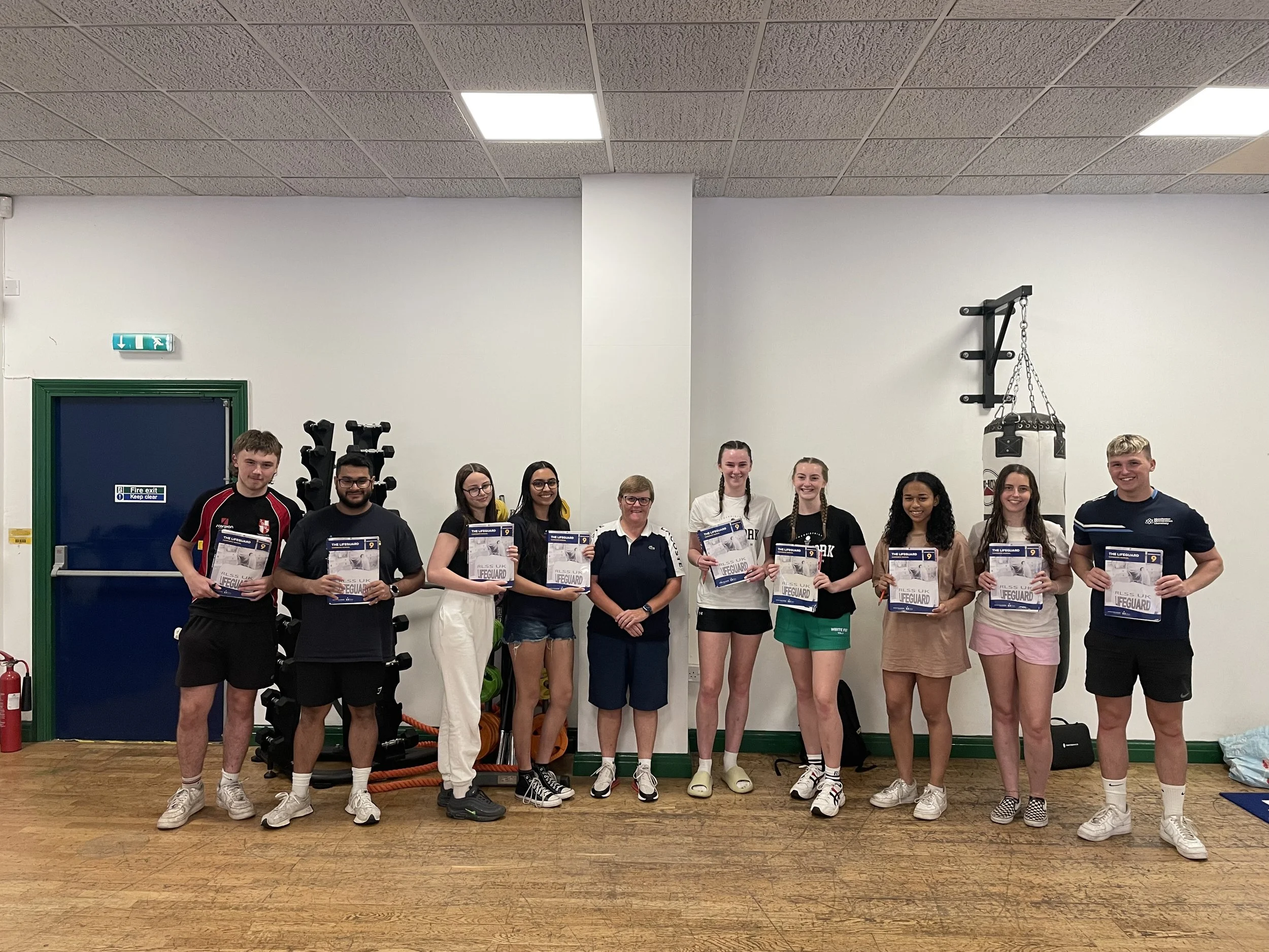 A group of ten young people and one adult standing indoors, holding certificates, in a fitness or gym setting.