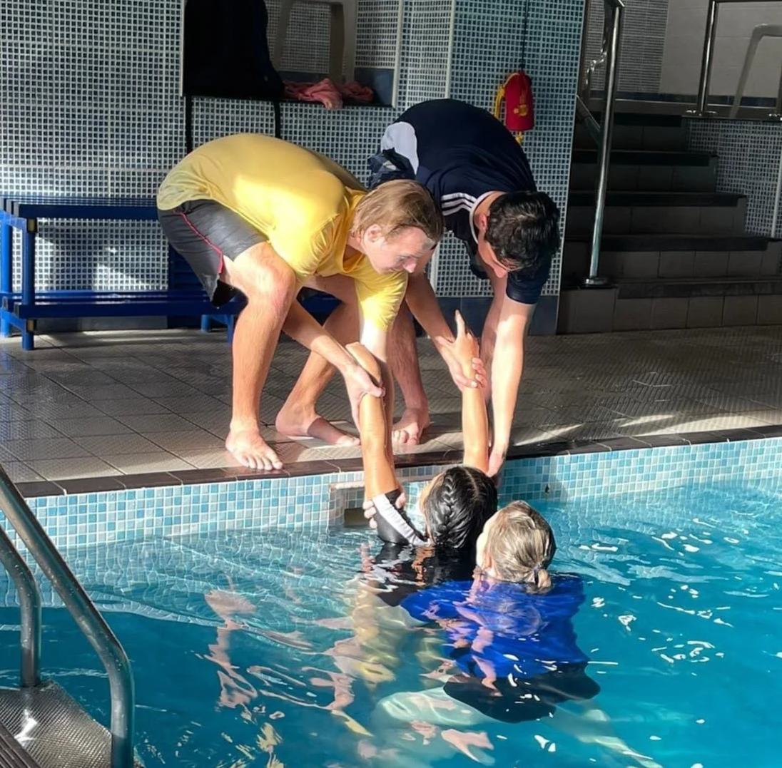 Two adults and two children at an indoor swimming pool, with the children in the water and the adults helping them out of the pool.