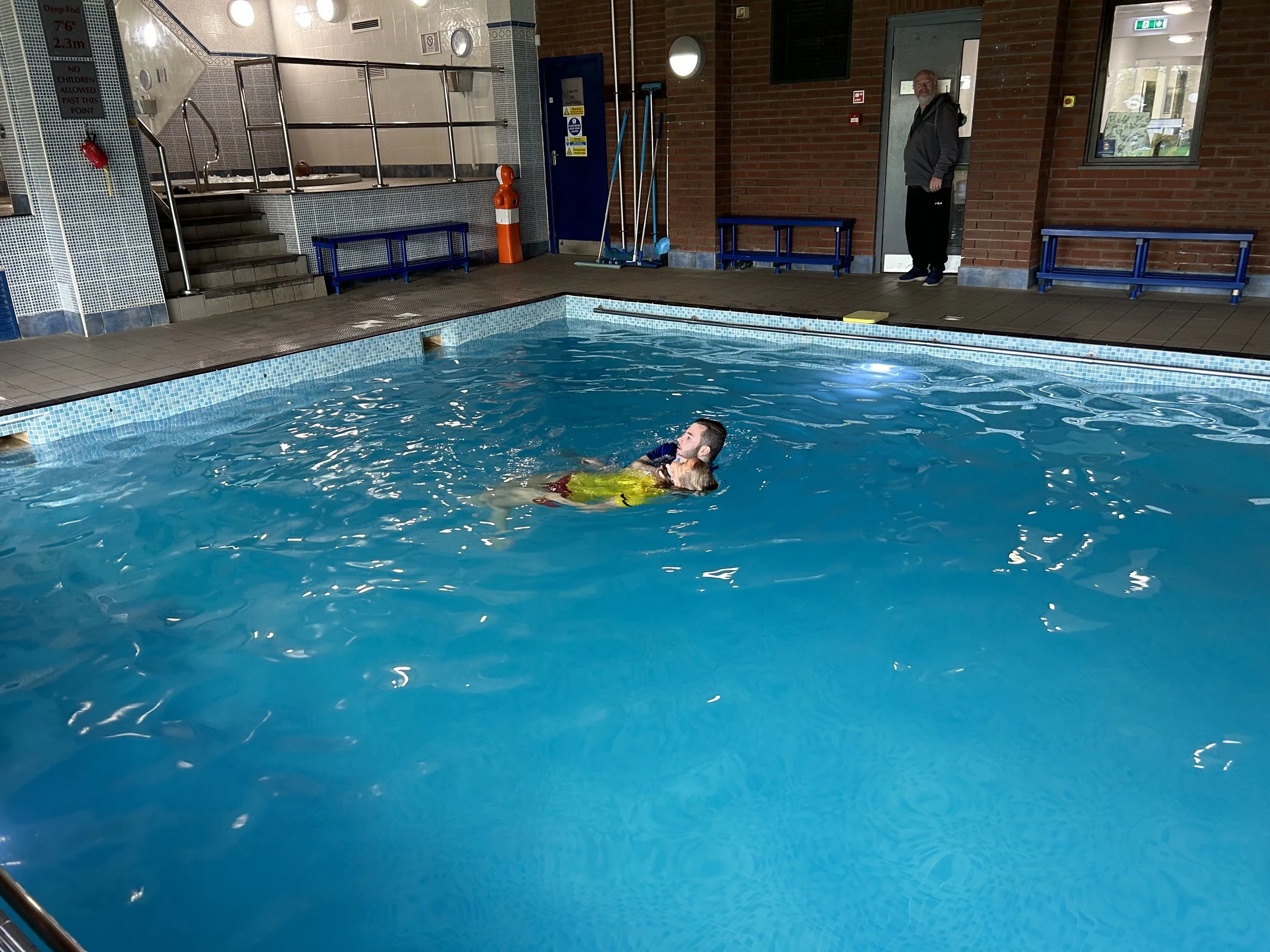An indoor swimming pool with a man and a child swimming together, the man holding the child in the water. In the background, a man stands near a brick wall and looks toward the pool. The pool area has railings, benches, and cleaning equipment, with b