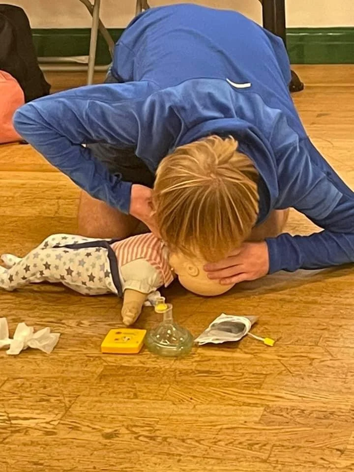 Adult practicing CPR on a doll on a wooden floor, surrounded by medical supplies including a mask and a yellow box.