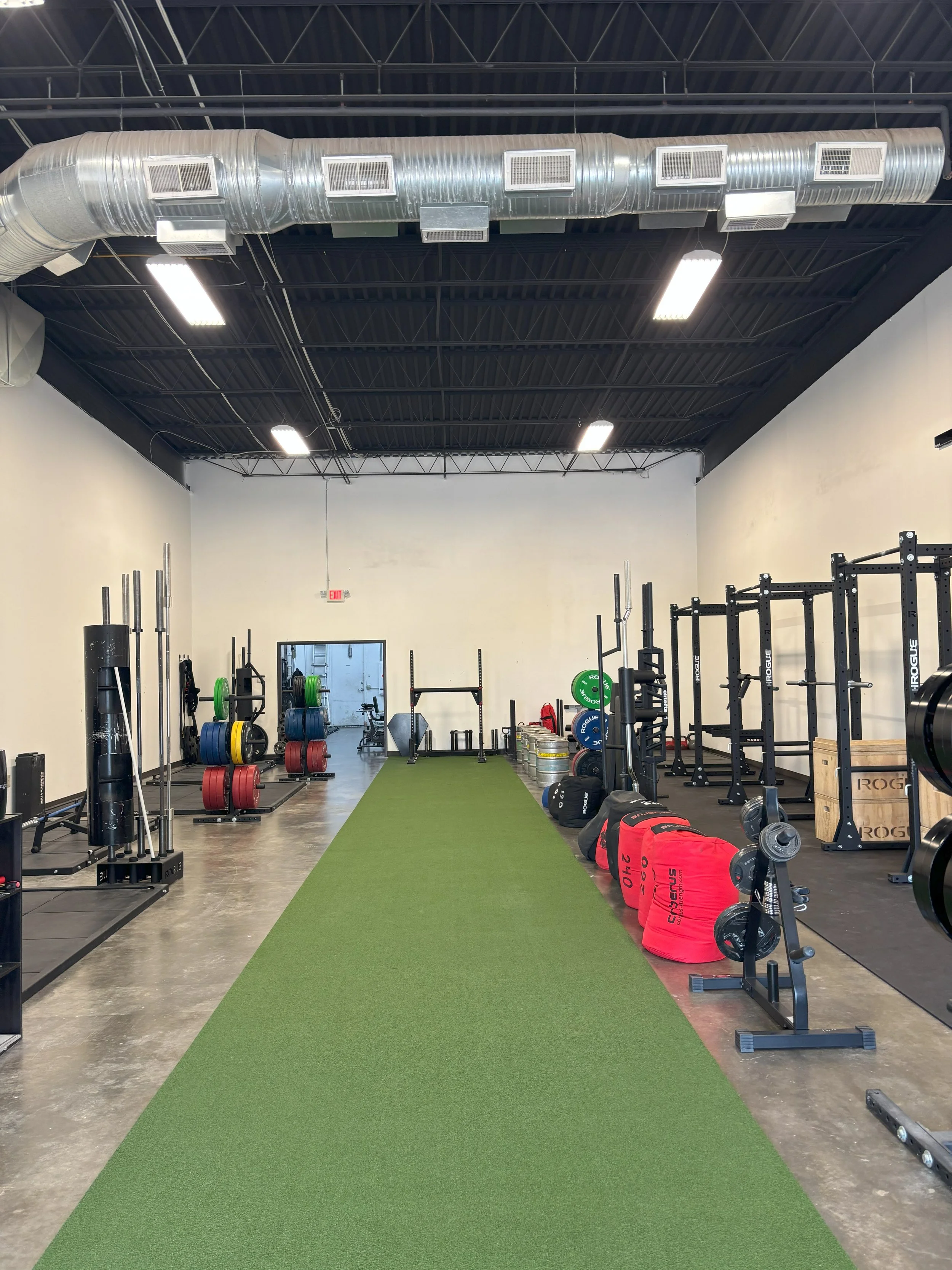 Empty indoor gym with weightlifting equipment, colorful weight plates, red sleds, and a green running strip on a concrete floor.