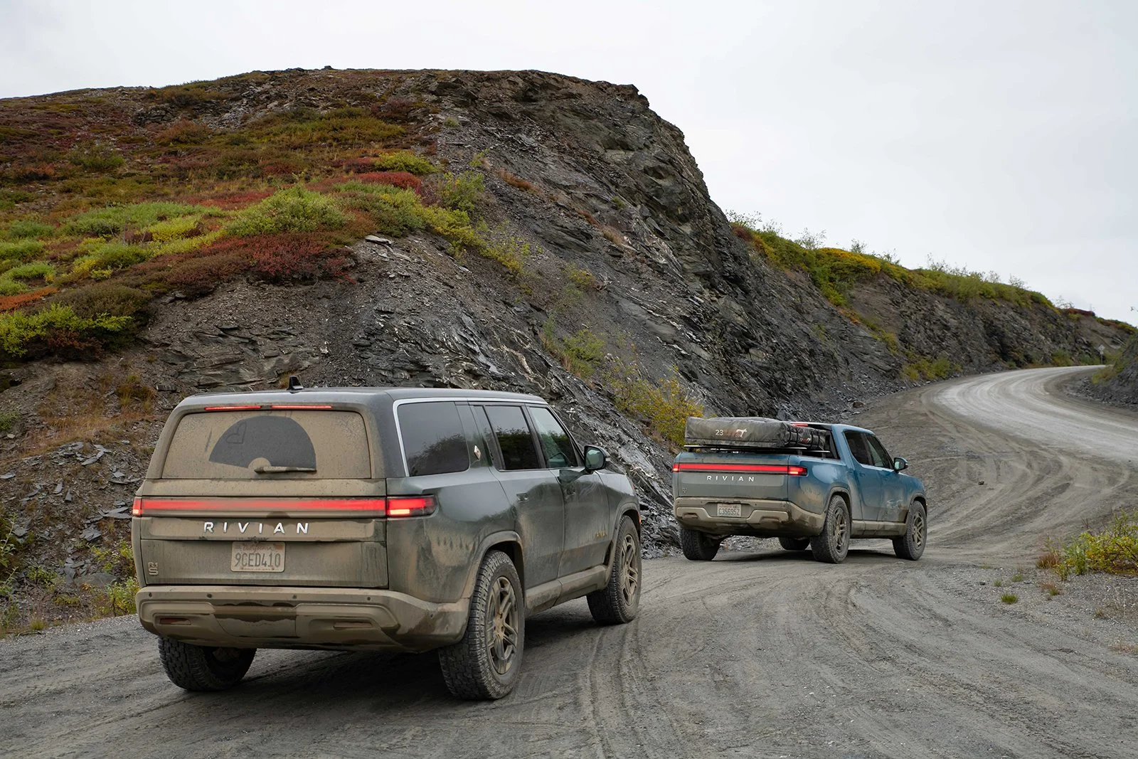 Two Rivian electric vehicles drive a muddy road on an overcast day
