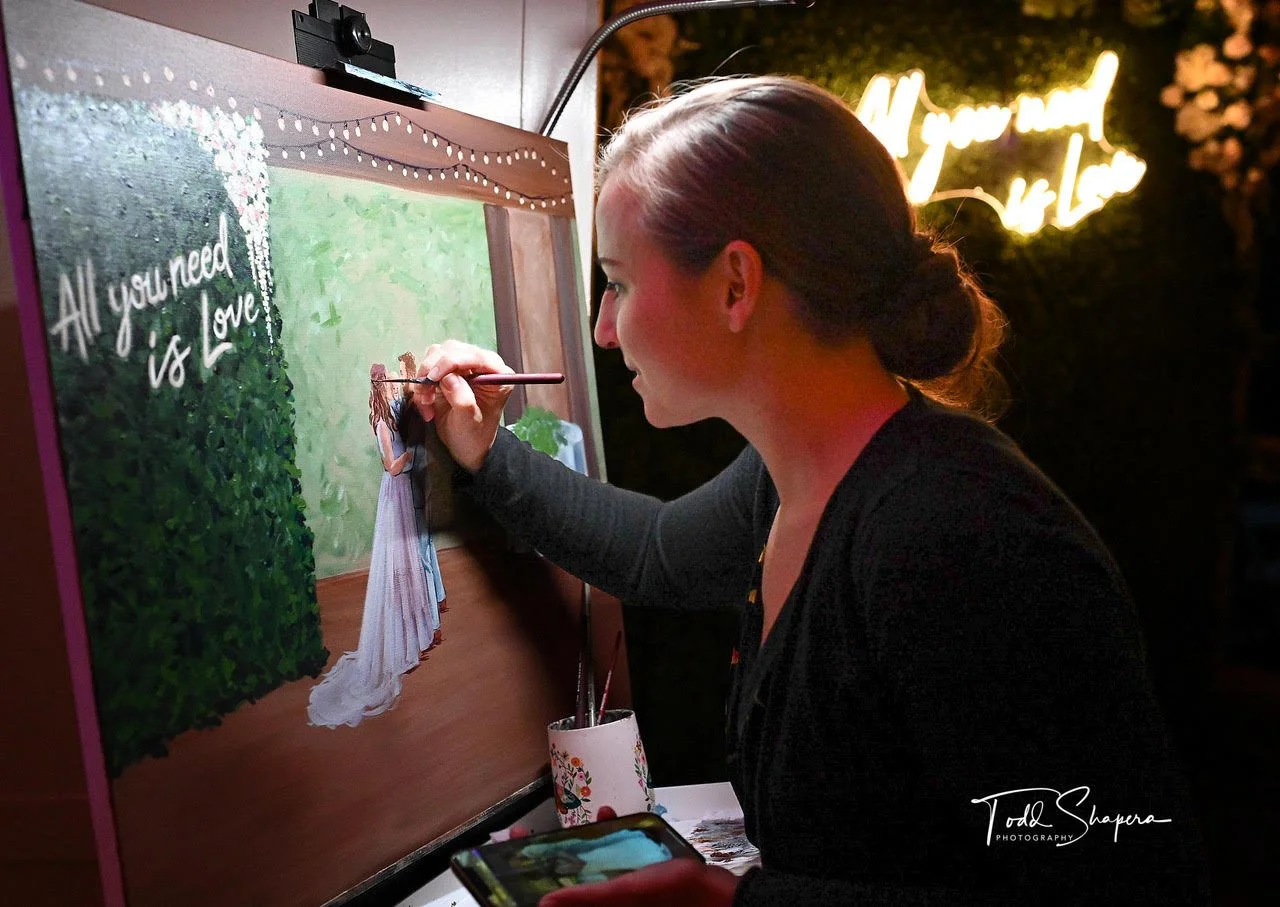 A woman with her hair tied in a bun is painting a wedding photo on a canvas at a celebration. The painting depicts a bride in a white wedding dress. The canvas has the words "All you need is Love!" written on it. In the background, there is a lit sign that reads "If you me" and some floral decorations.