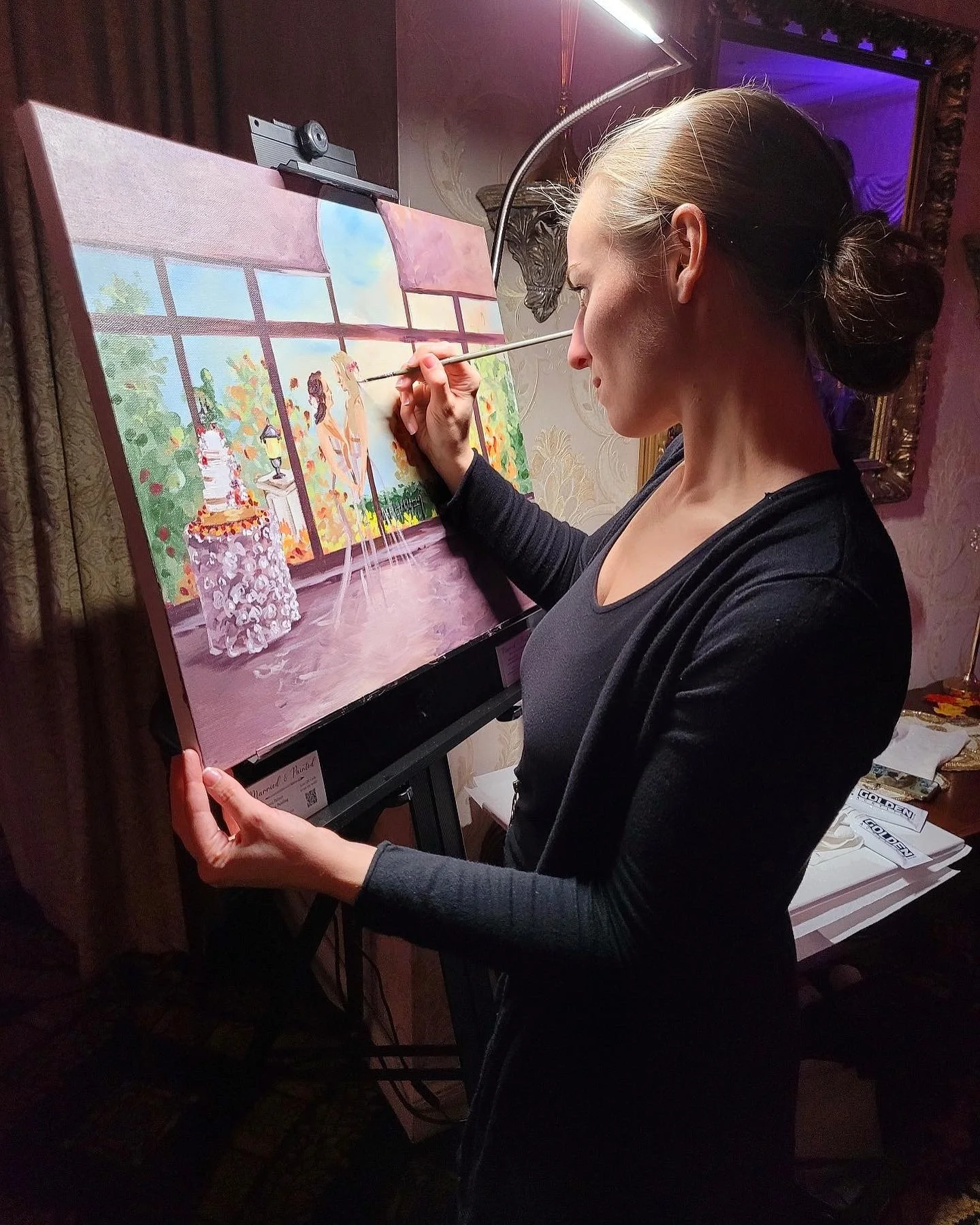 A woman painting on a canvas with a view of a house and garden scene, standing indoors with ornate wallpaper and framed decorations.