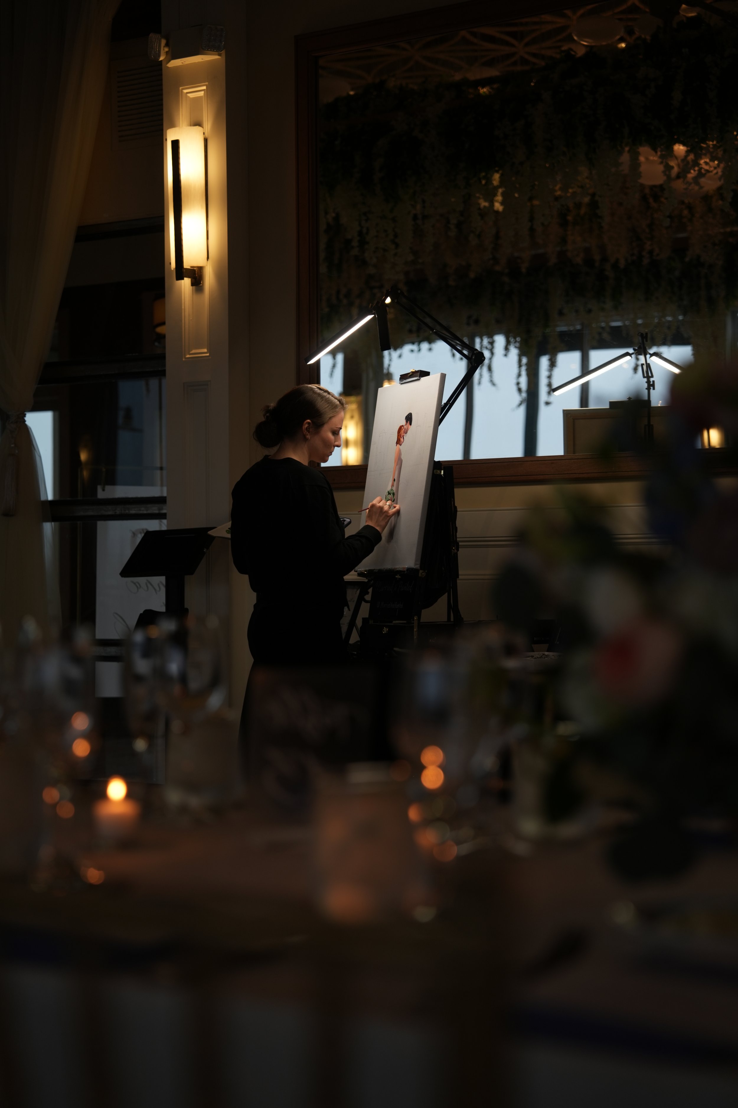 A woman is painting a portrait on a canvas in a dimly lit indoor space with large windows, greenery outside, and tables with candles and glassware in the foreground.