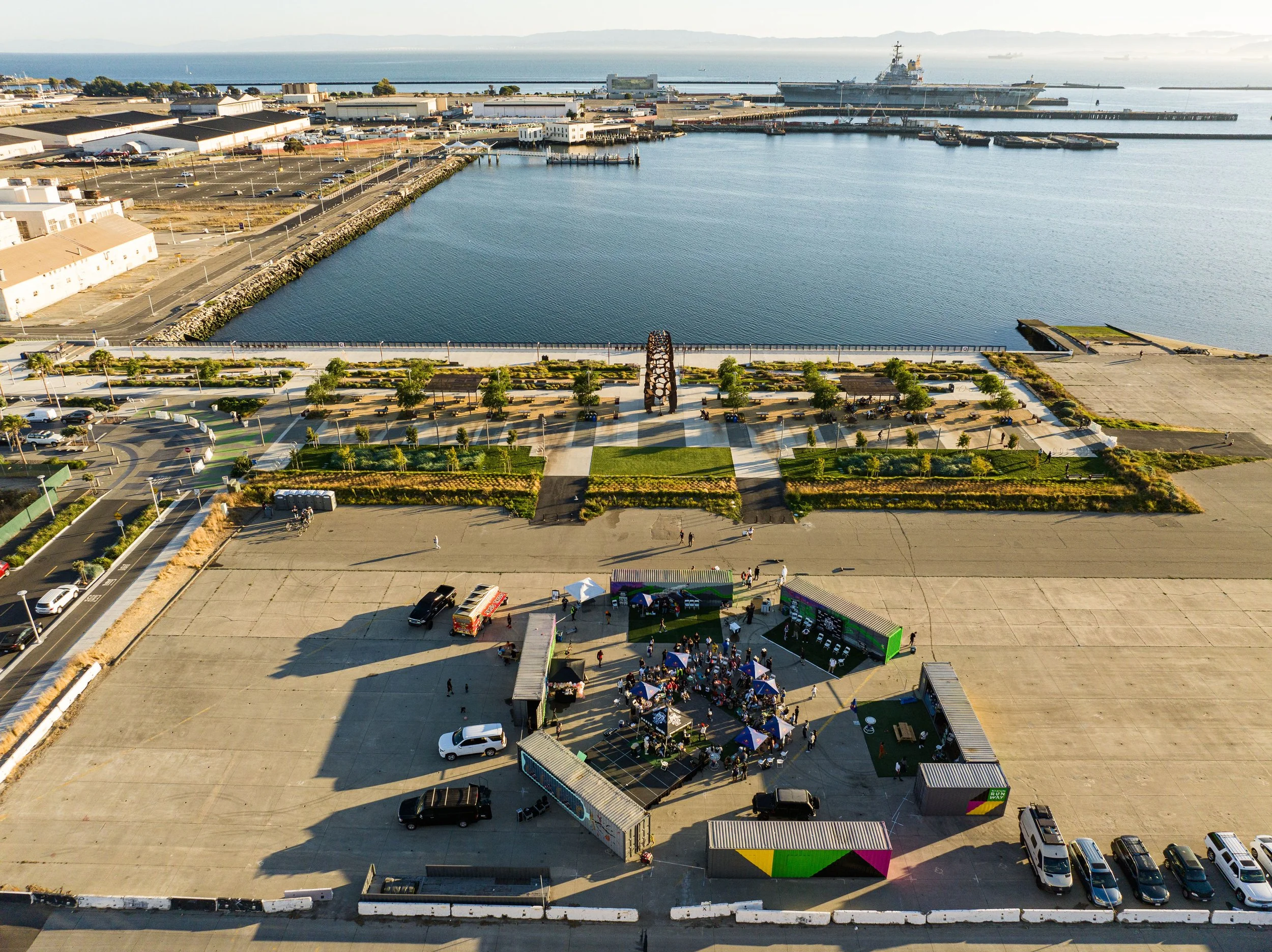 An aerial view of a waterfront area with a large parking lot, a small event with tents and people, and a park with trees and benches near the water, with ships docked in the harbor in the background.
