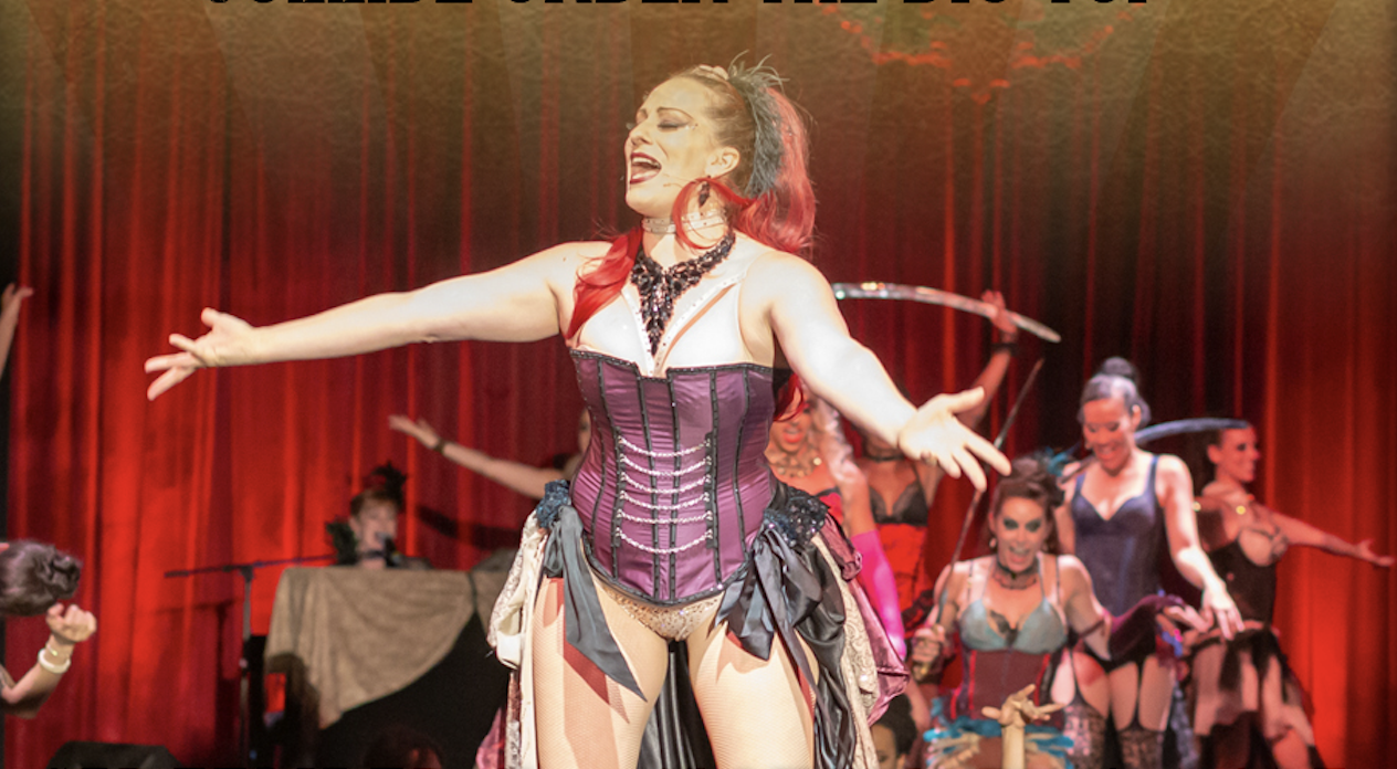 Performer on stage wearing a purple and white corset, with red and black hair, singing with arms outstretched, surrounded by other performers in colorful costumes and fishnets in front of a red curtain.