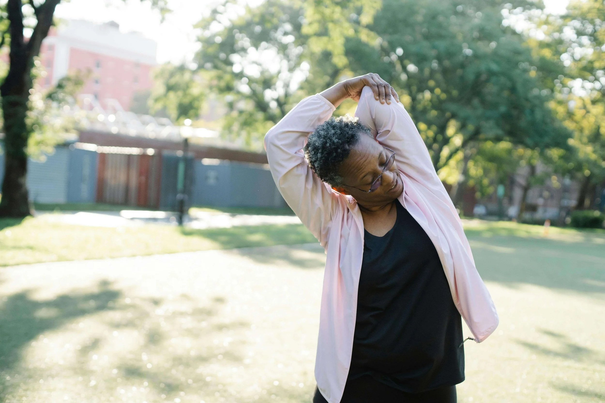 Older woman wearing a pink active jacket stretching her arms overhead outdoors, illustrating release and self-care in trauma therapy for emotional parentification in Falls Church, VA.