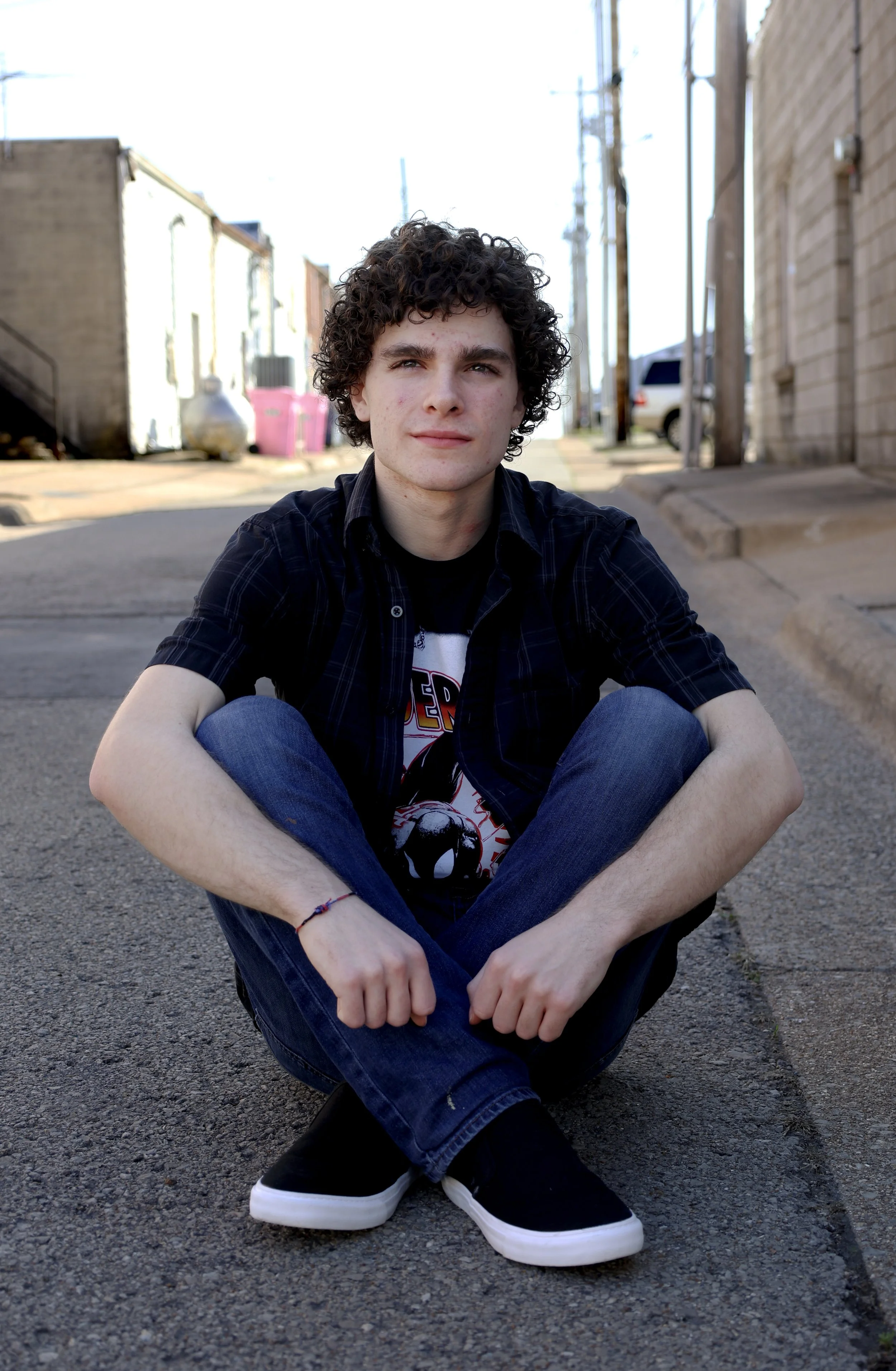 A young man with curly hair sits on the pavement in an urban alleyway, wearing a black and white graphic t-shirt, a dark plaid shirt, jeans, and sneakers with black and white soles. There are buildings, power lines, and parked vehicles in the backgro