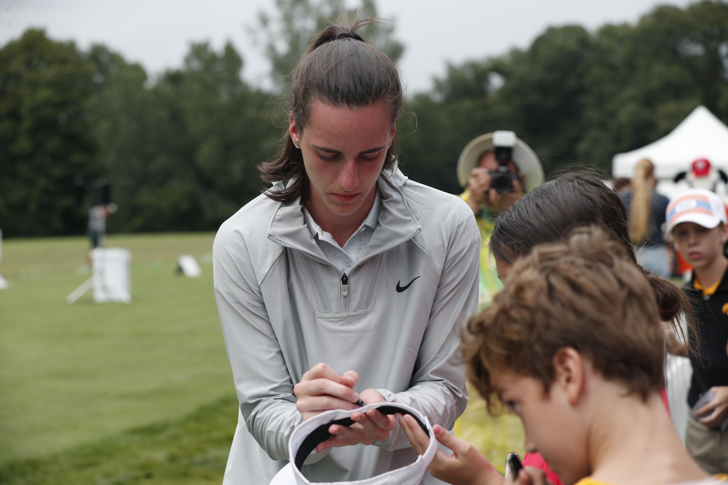A young woman in a gray Nike jacket with brown hair tied in a ponytail is signing an autograph for a young girl at an outdoor event. In the background, there are other children, a person with a camera, and a white tent, with trees and an overcast sky