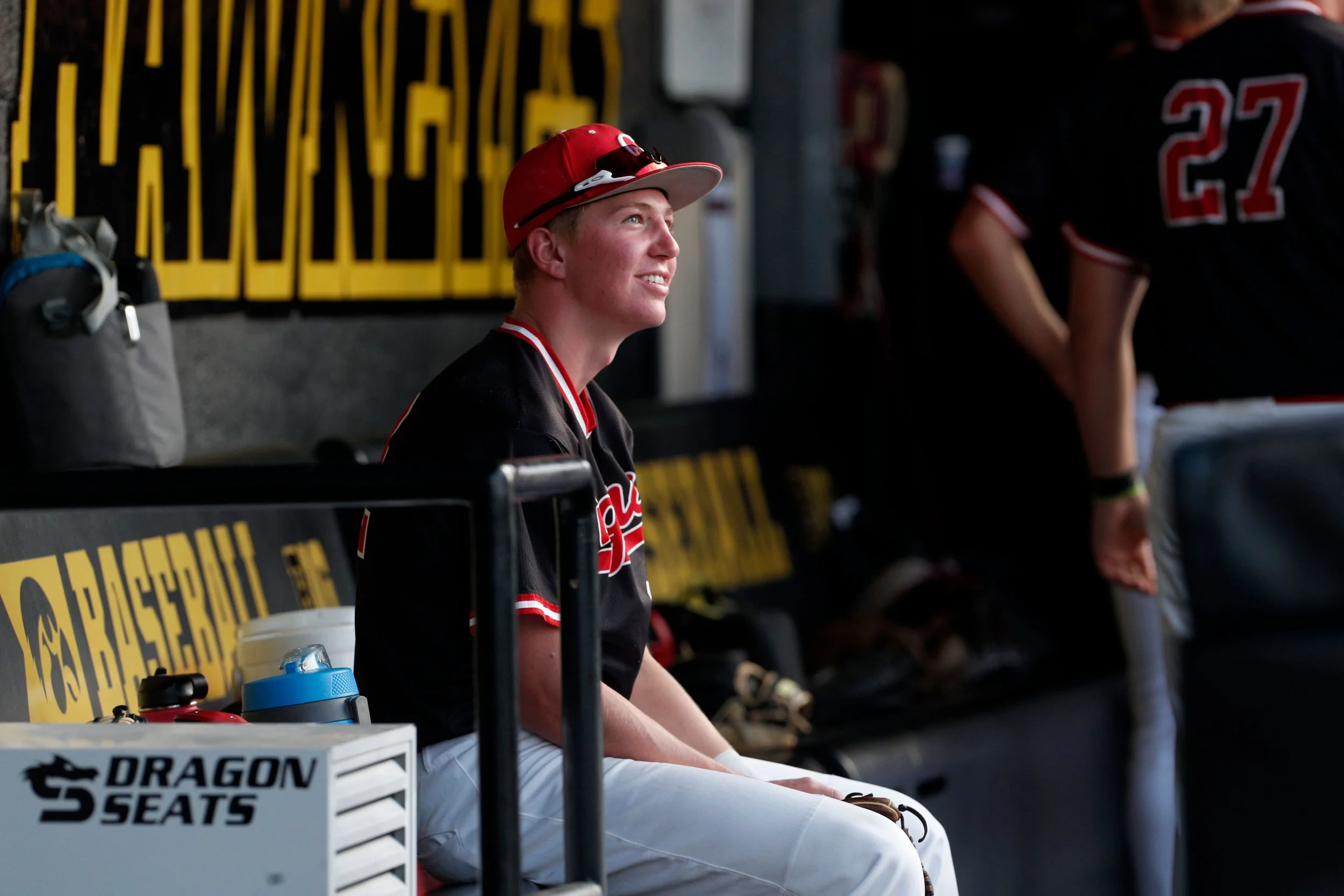 A young man wearing a red baseball cap, sunglasses, and a black and red sports jersey sitting in a garage or pit area, smiling and looking upward. There is a yellow safety barrier and sports equipment around him.