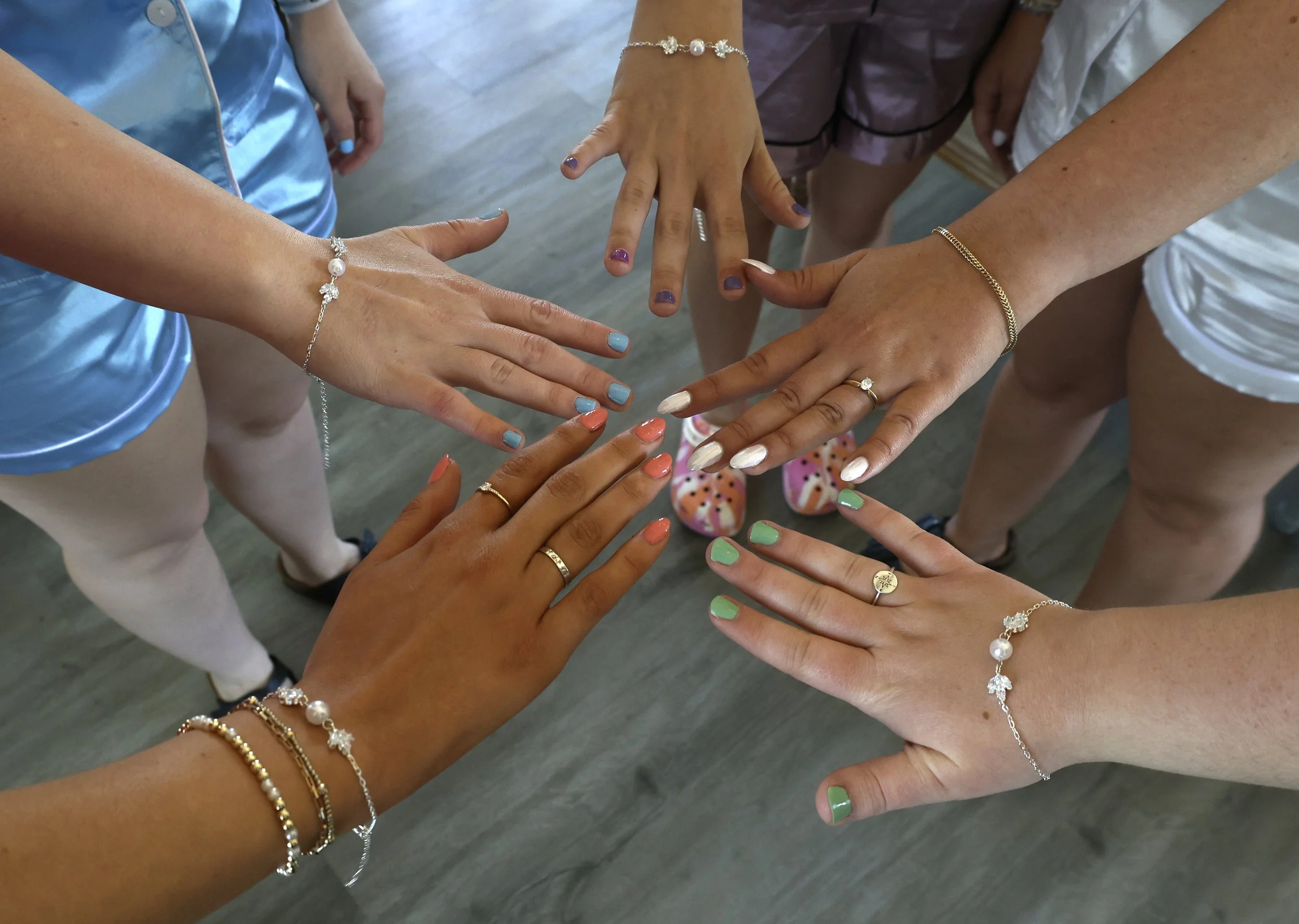 Multiple hands with colorful nail polish showing jewelry such as bracelets and rings, arranged in a circle on a wooden floor.