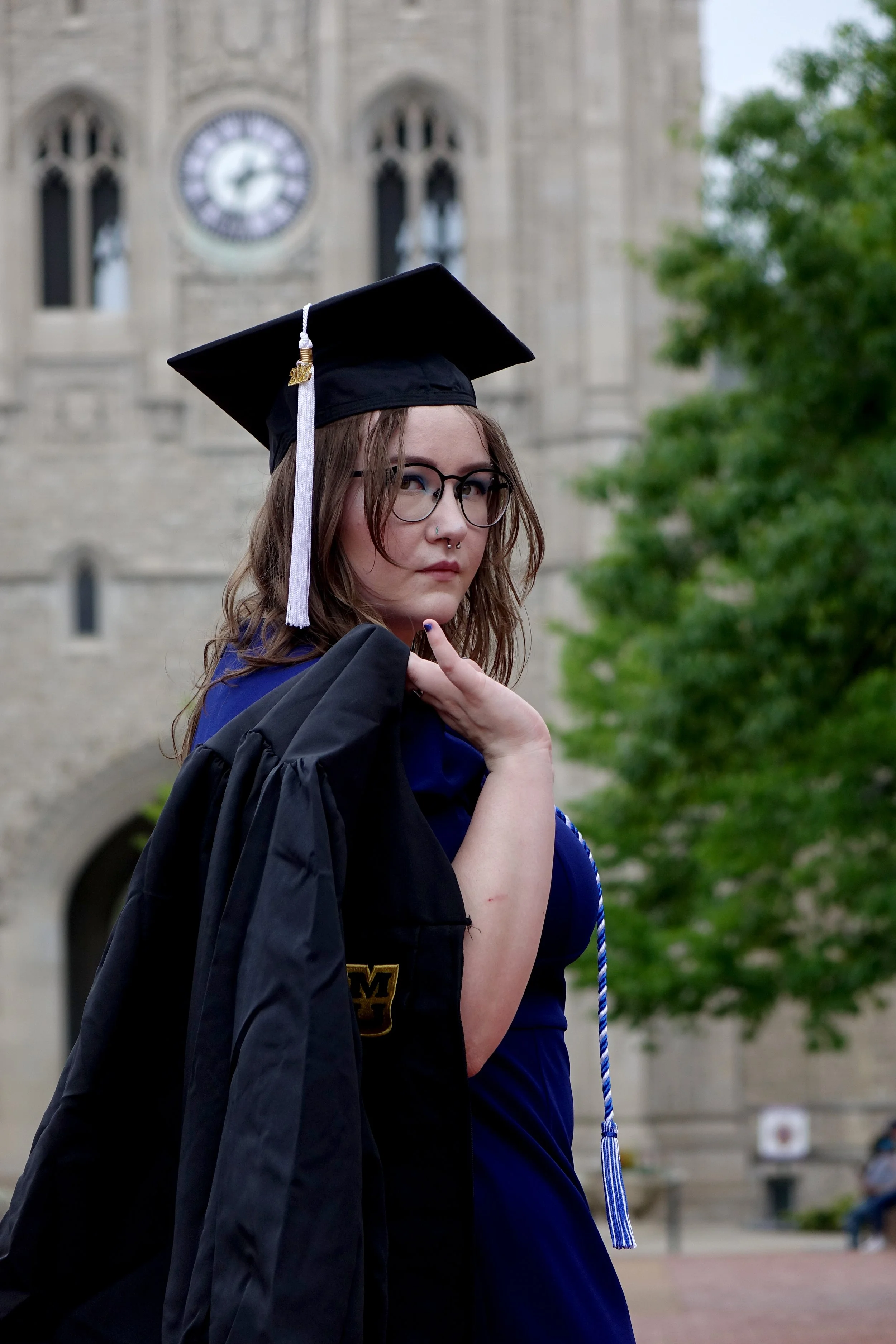 Young woman in graduation cap and gown standing outdoors in front of a stone building with a clock tower.