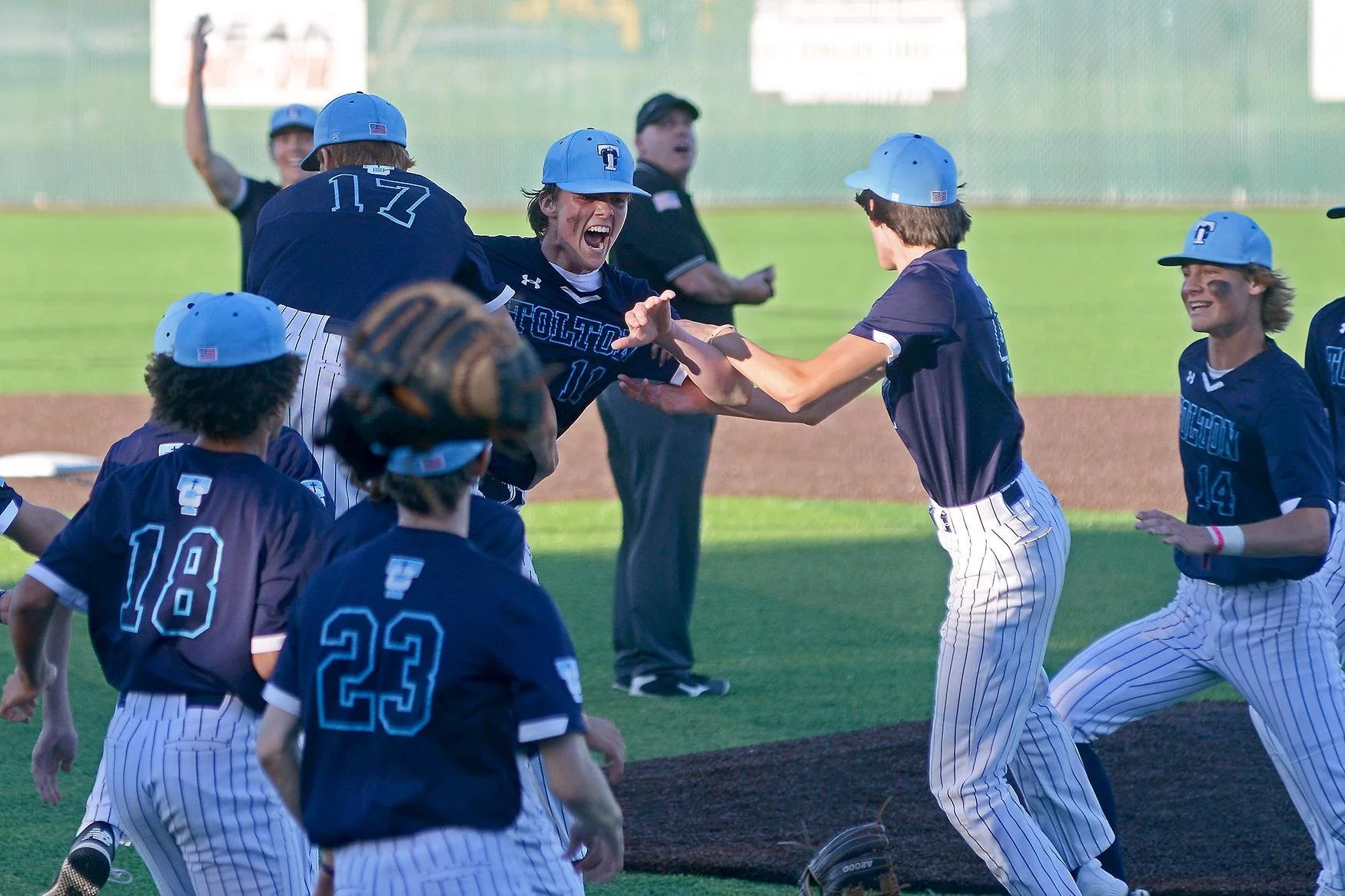 A group of young baseball players wearing blue jerseys and striped pants celebrating on the field. They are smiling, high-fiving, and running together with a coach or umpire in the background.