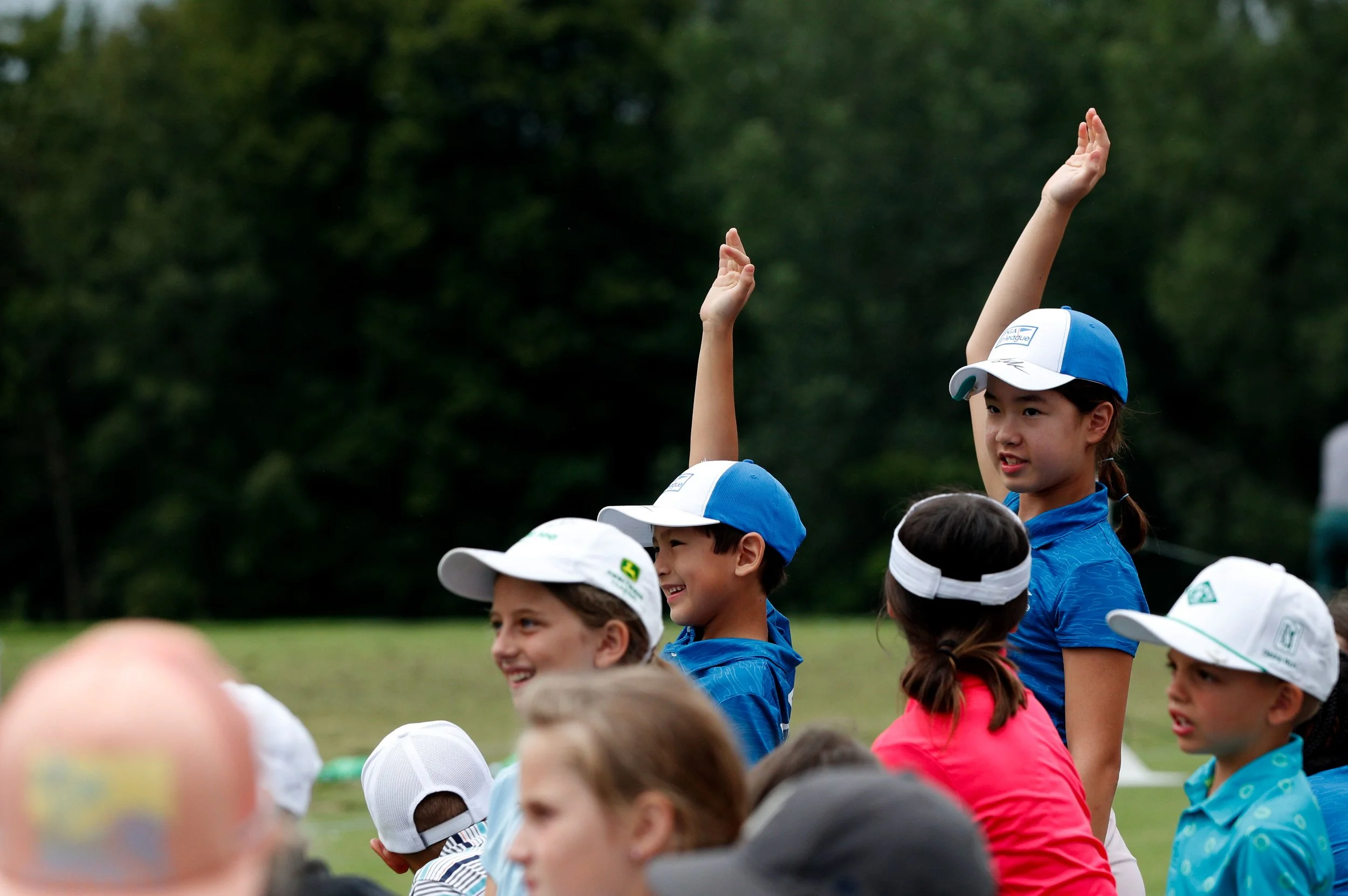Children gathered outdoors, with one girl raising her hand, wearing a blue shirt and a blue and white cap, among others who are smiling and looking in various directions.