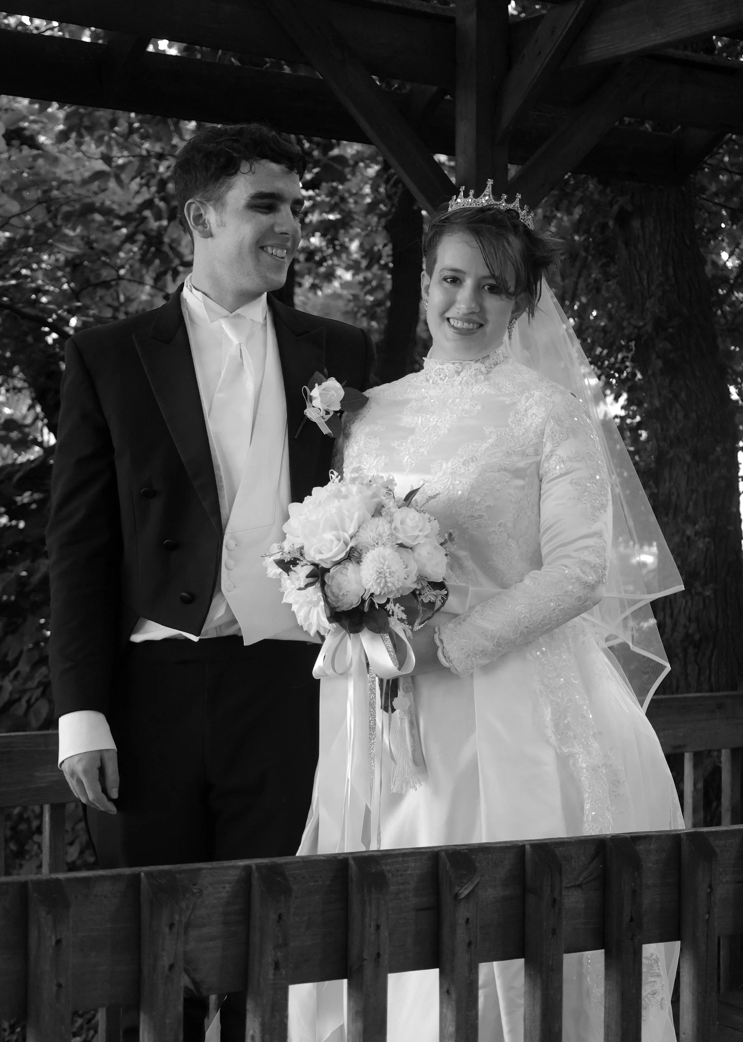 Black and white photo of a smiling man in a tuxedo and a smiling woman in a wedding dress wearing a crown, holding a bouquet of flowers, standing outside under a wooden structure with trees in the background.