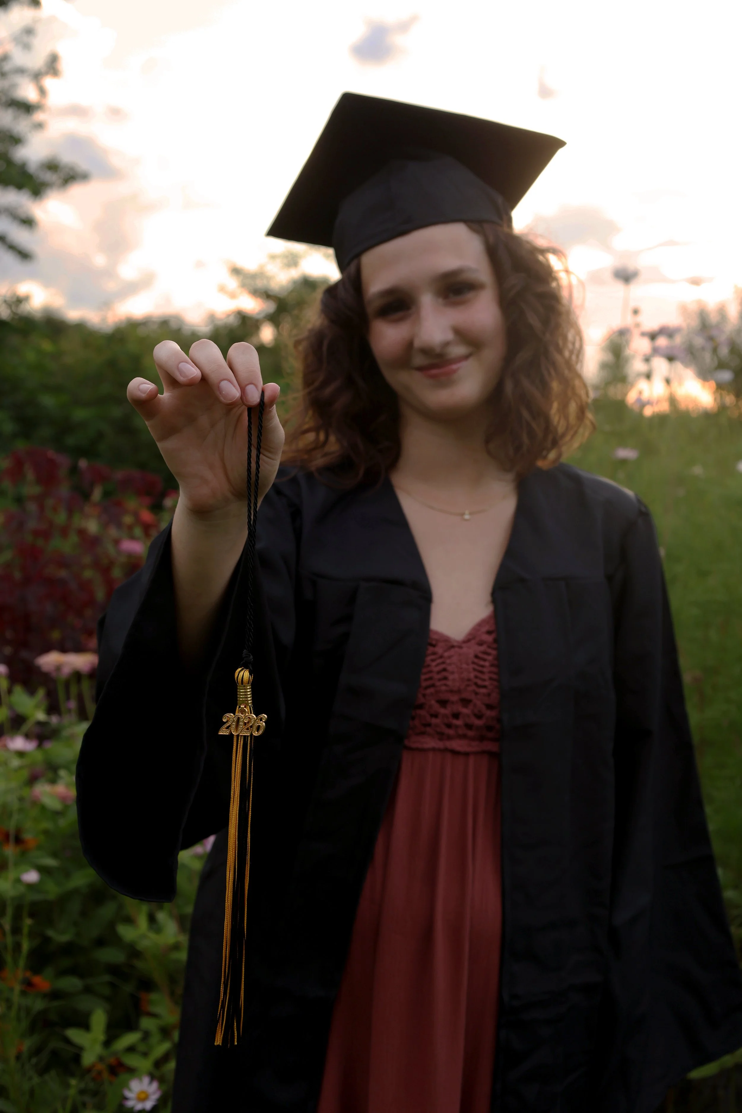 A woman in a graduation gown and cap holding a necklace with a '2026' charm outside during sunset.