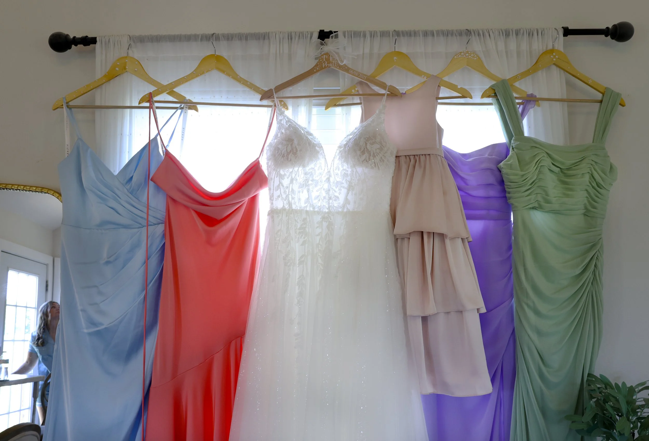 Colorful dresses hanging on a black rod in front of a window with white curtains. The dresses are in various pastel colors including blue, red, white, pink, purple, and green, each with different style and fabric details.