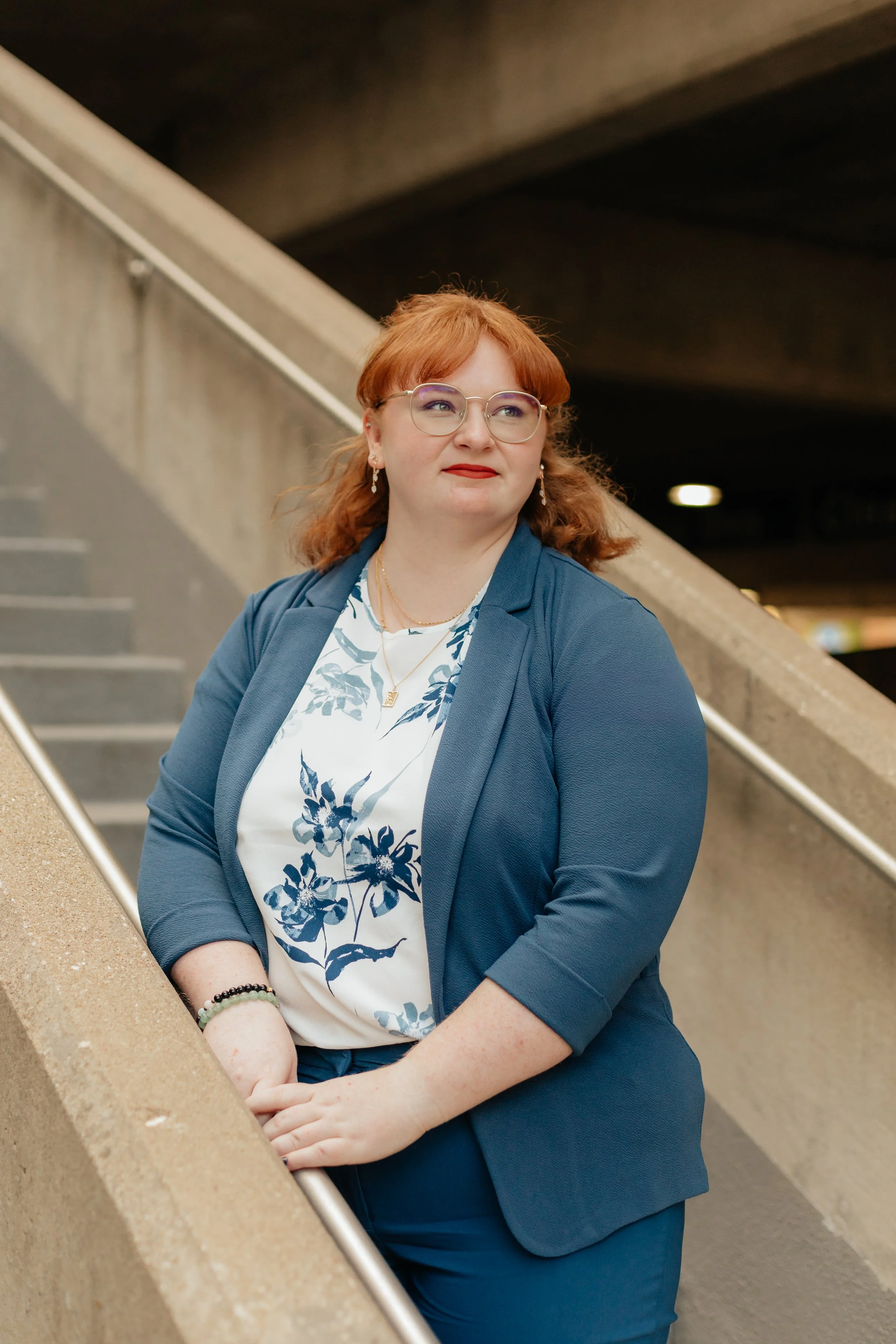 A woman with red hair, glasses, and red lipstick standing on a staircase in an indoor setting, wearing a blue blazer and a white blouse with a blue floral pattern.