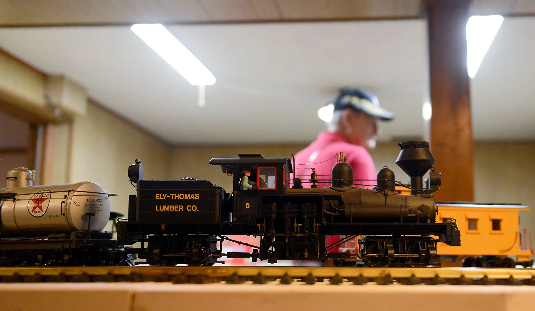 Close-up of a detailed model train engine labeled 'Ely-Thomas Lumber Co.' on a track, with a person in the background wearing a pink shirt and cap, observing the display indoors.