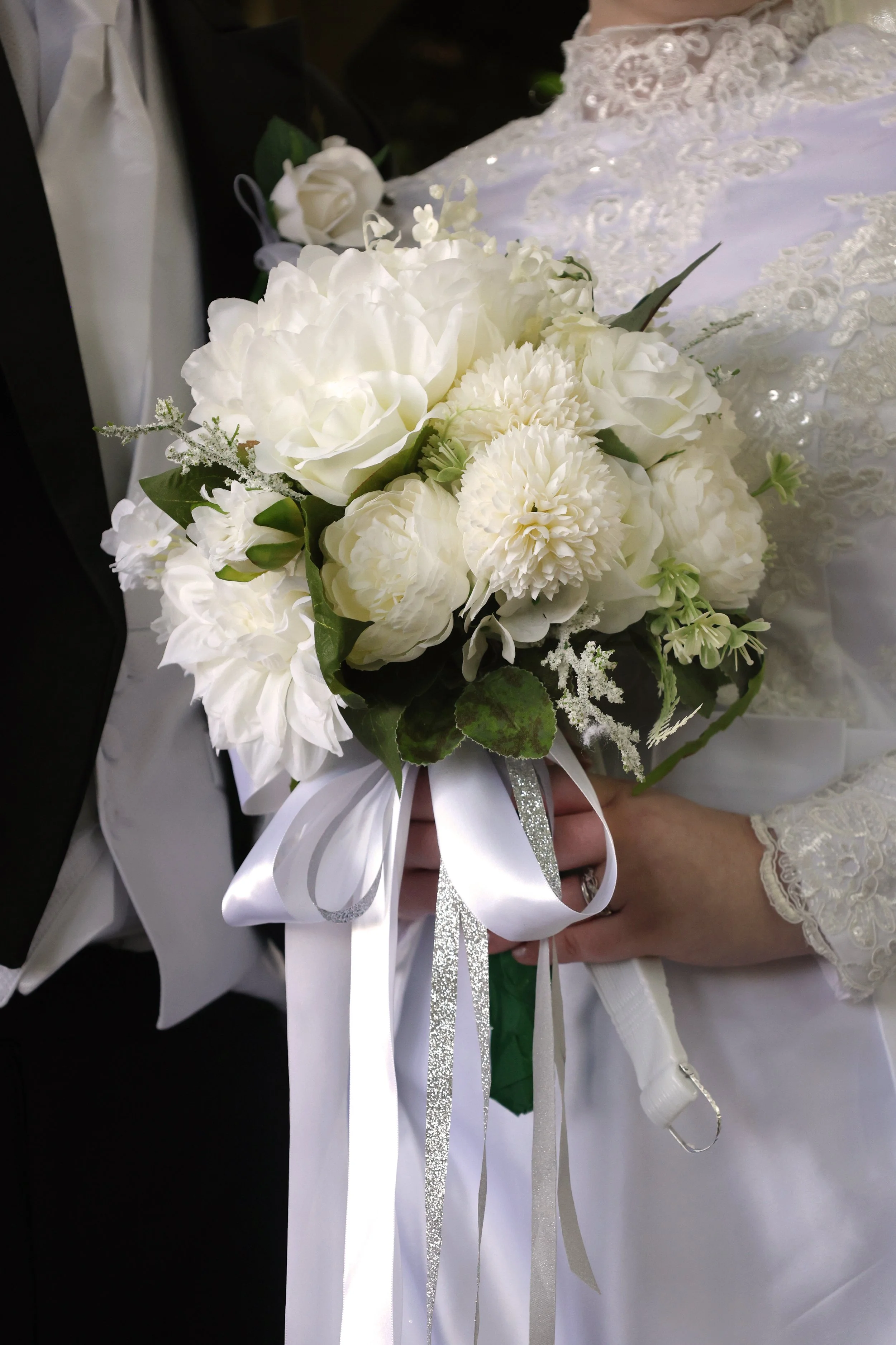 A bride holding a bouquet of white roses, carnations, and greenery, with ribbon accents, in a wedding dress with lace embroidery.