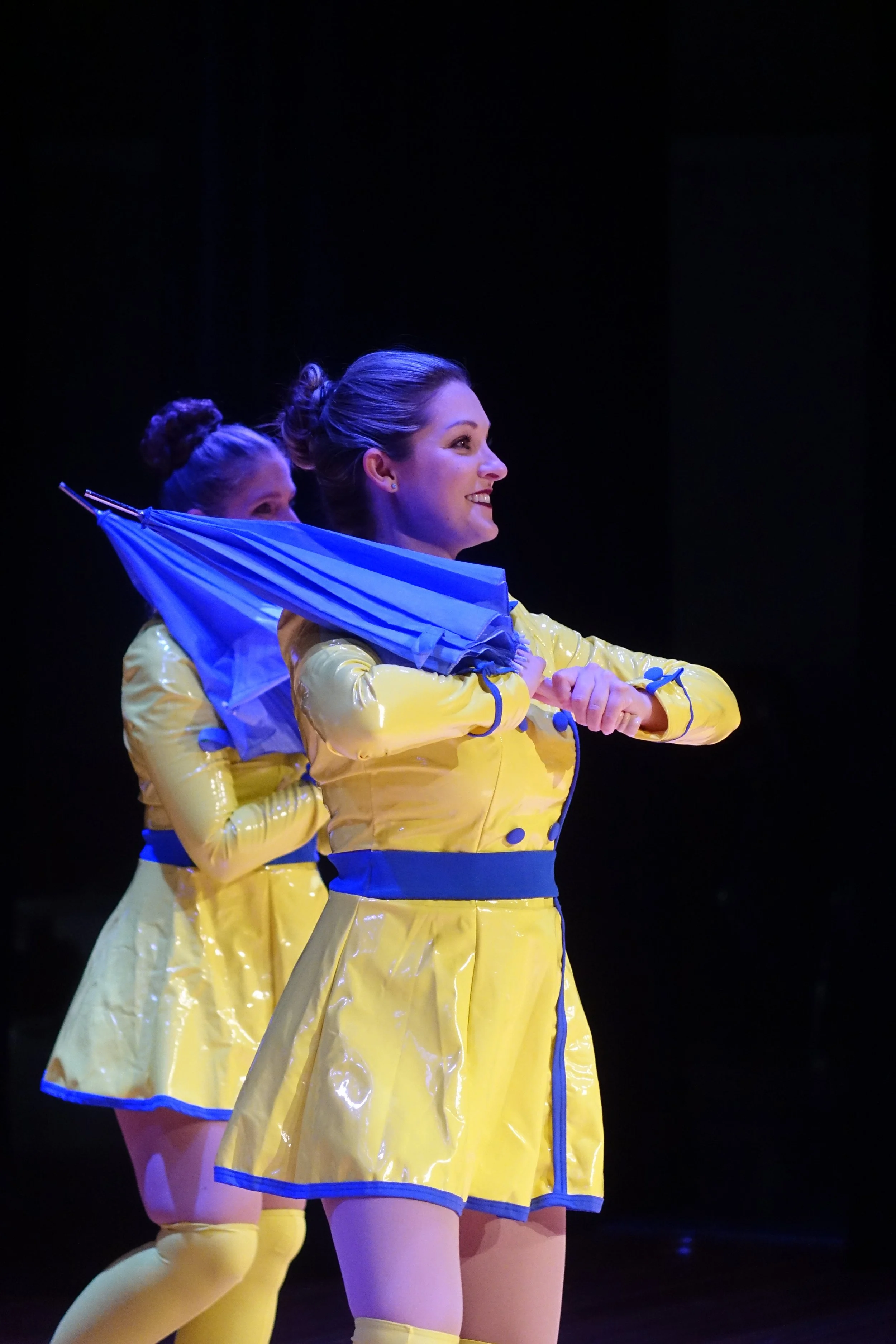 Two women dressed in shiny yellow costumes and yellow thigh-high stockings on stage, holding blue umbrellas, performing in unison with one smiling woman in the foreground and another woman partially visible in the background.