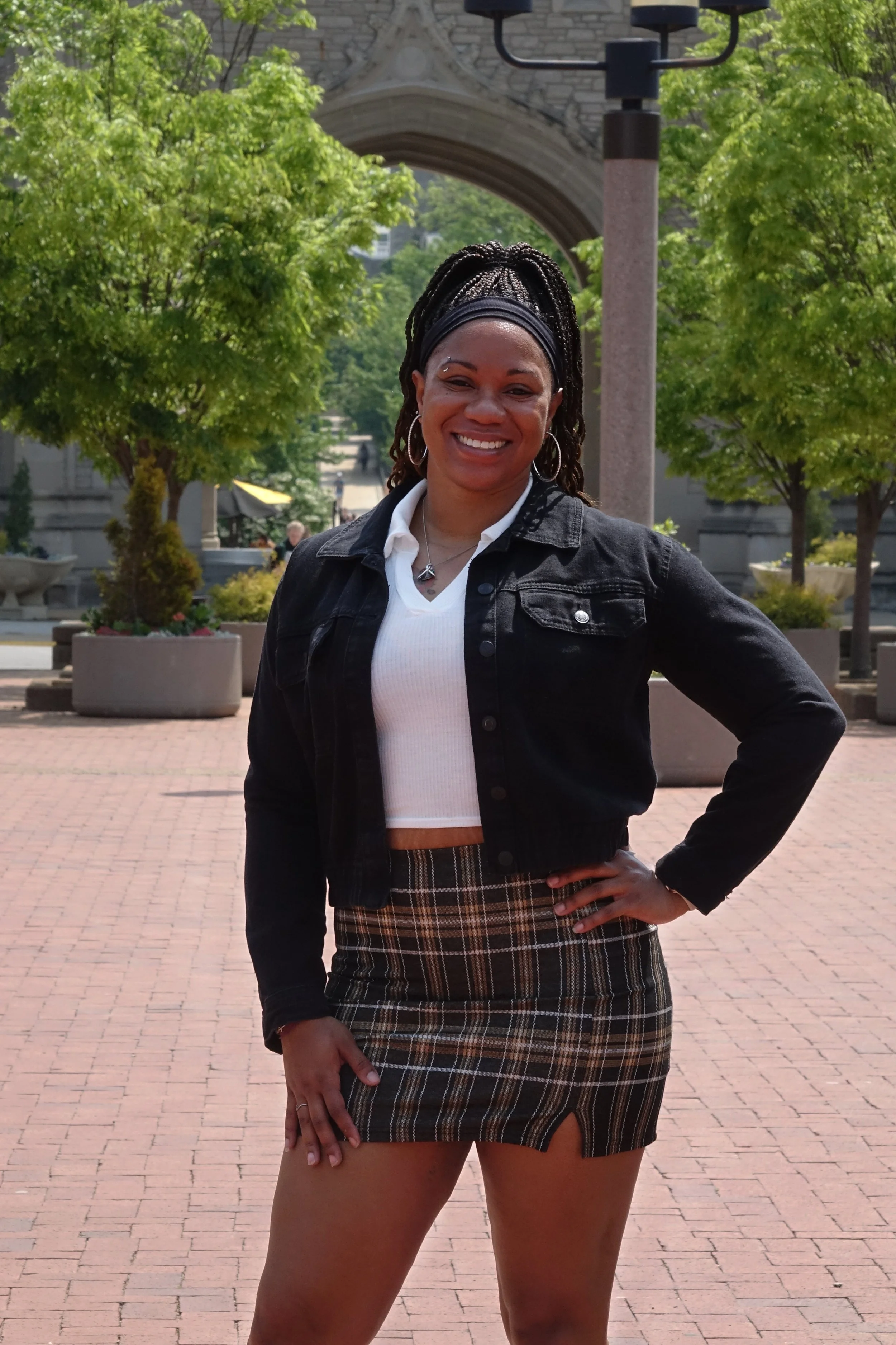 A woman standing outdoors in a park with trees and a stone archway in the background. She is smiling, with black braids, hoop earrings, and wearing a black jacket over a white top and a plaid skirt.