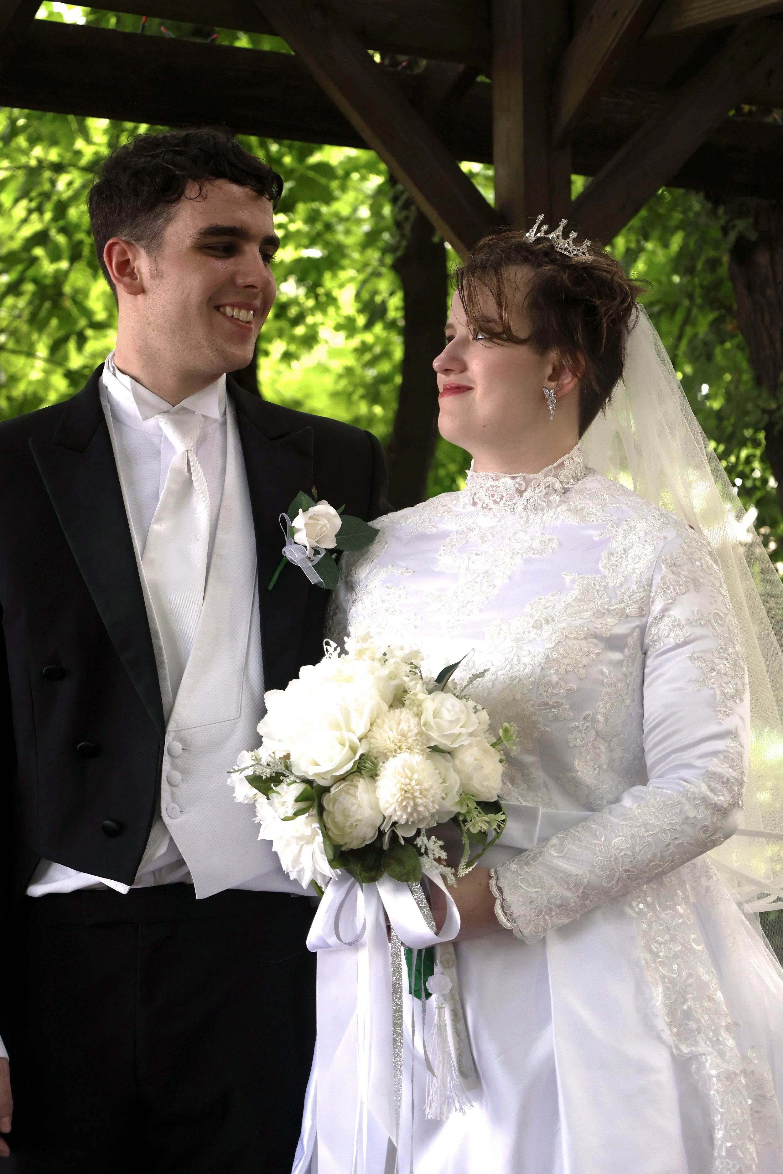 A bride and groom smiling at each other during their wedding ceremony outdoors. The bride is wearing a white lace wedding dress, a veil, and a tiara, holding a bouquet of white flowers. The groom is dressed in a black tuxedo with a white shirt and ti