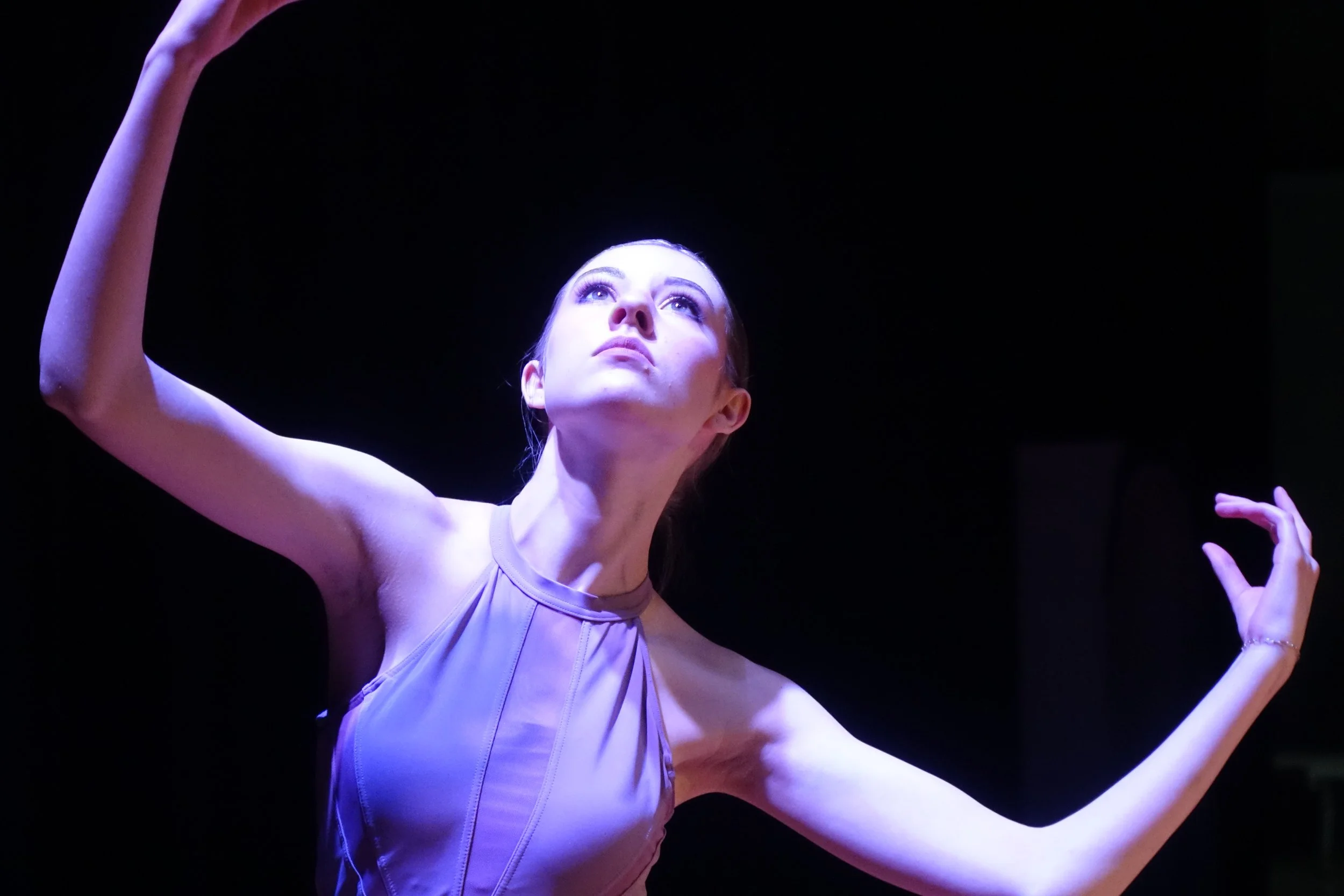 A female dancer performing on stage, with arms raised and head slightly tilted back, illuminated by stage lighting, black background.