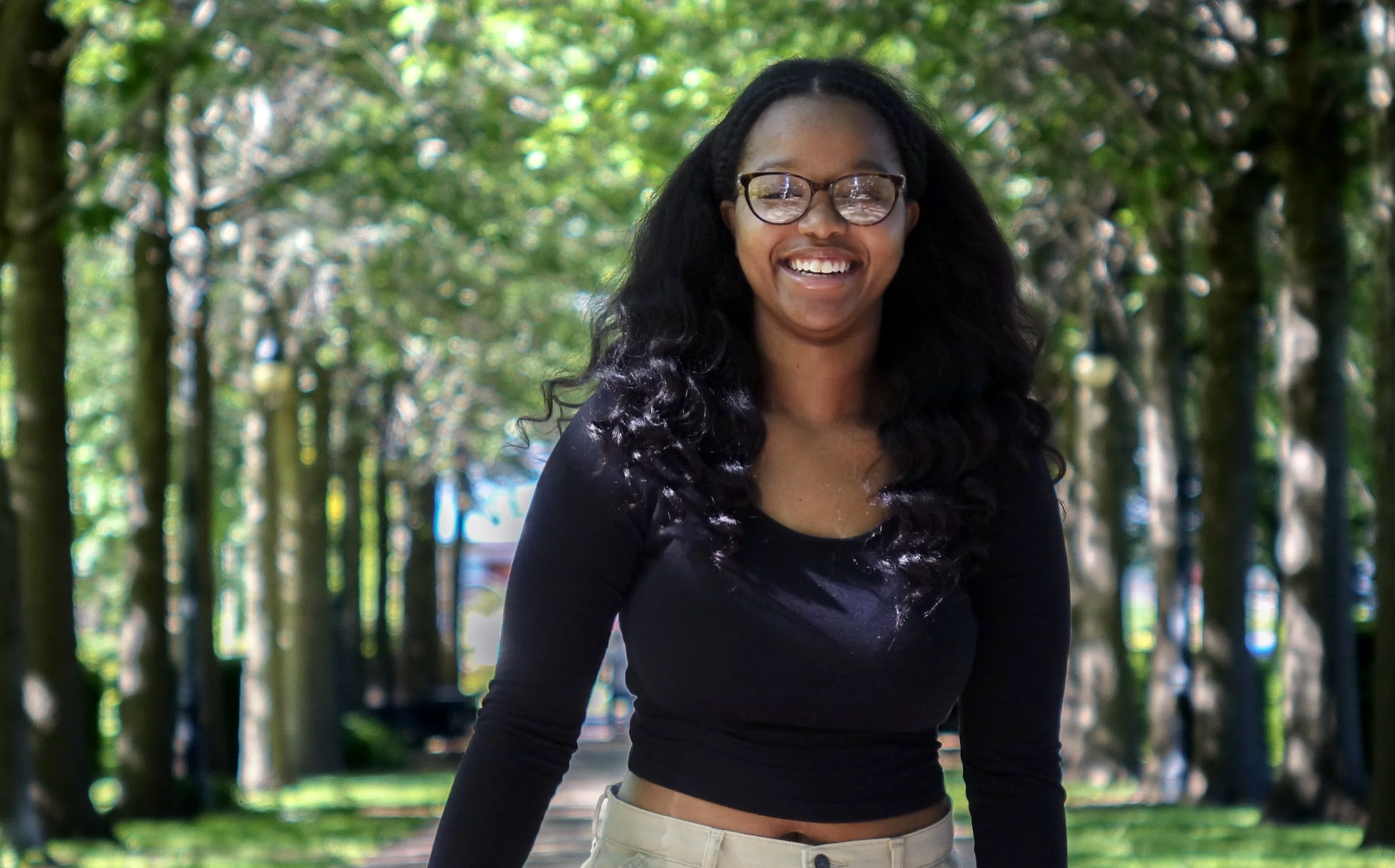 A smiling woman with glasses, long curly black hair, wearing a black top and beige pants, standing outdoors in a park with green trees in the background.
