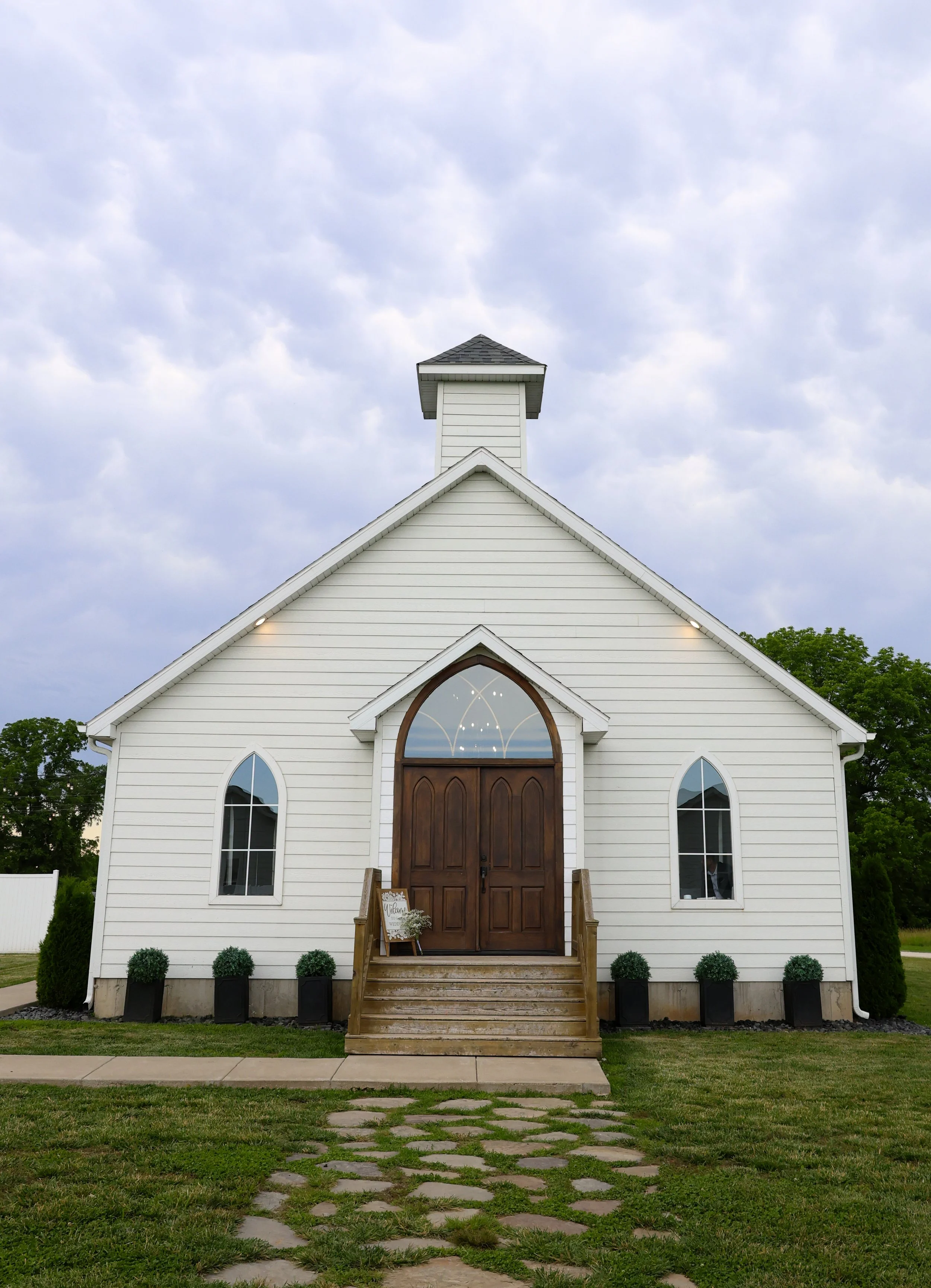 White church with wooden front door, arched windows, steps leading up to the entrance, and a steeple on the roof, set against a cloudy sky with patches of greenery.