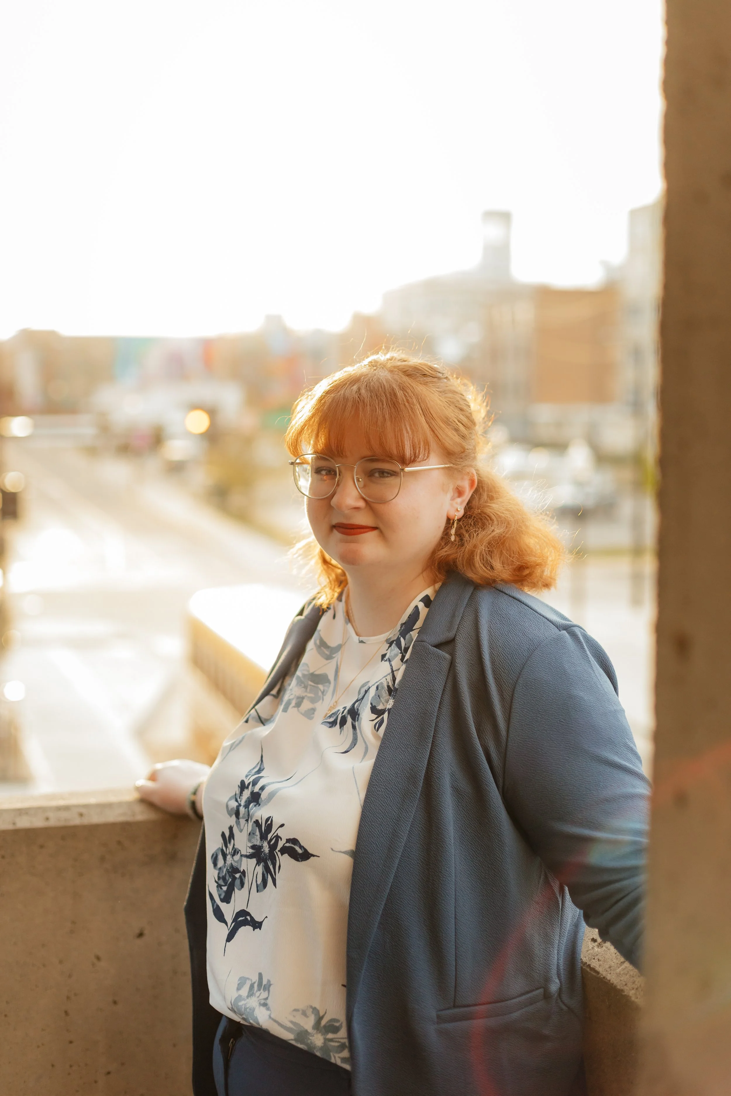A woman with red hair, glasses, and earrings standing outdoors on a sunny day, leaning against a concrete ledge, with buildings and a cityscape blurred in the background.