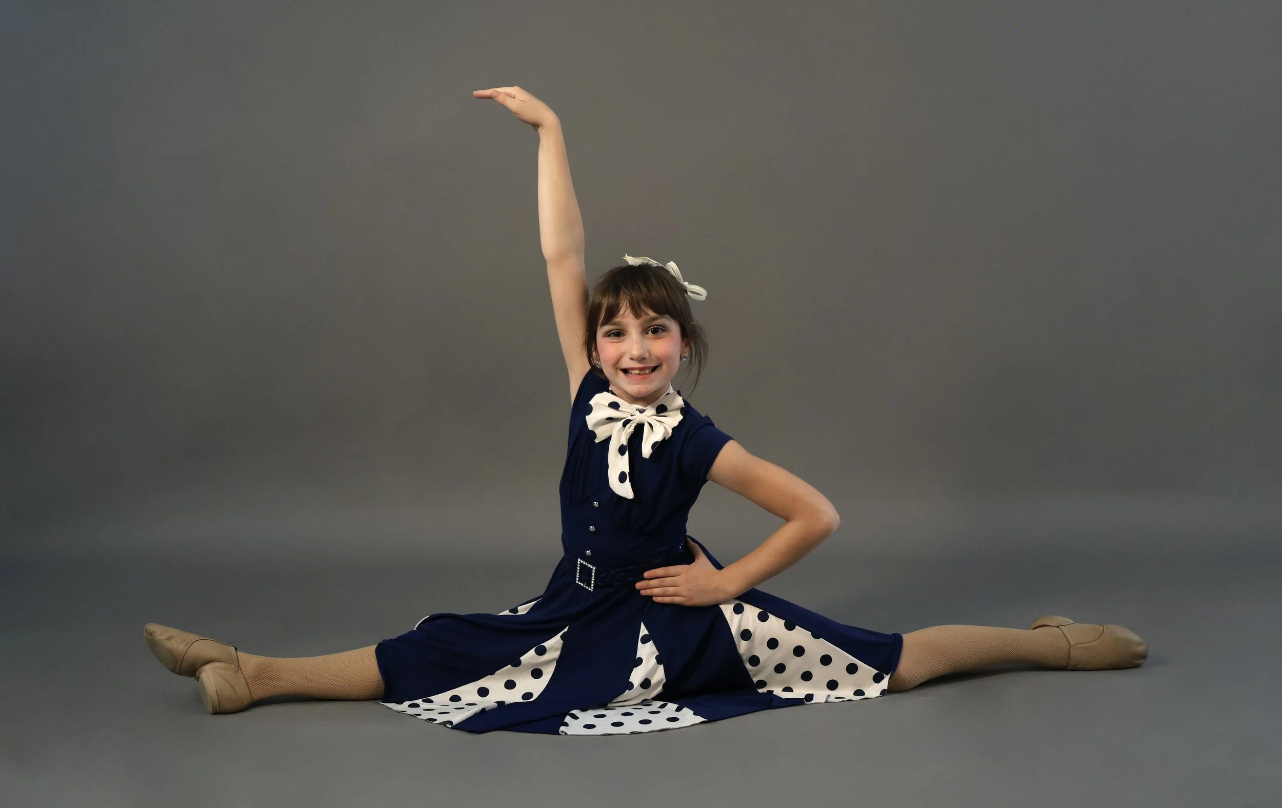 Young girl in a navy blue dress with polka dots, performing a split on a gray studio background, smiling and posing with one arm raised and the other on her waist.