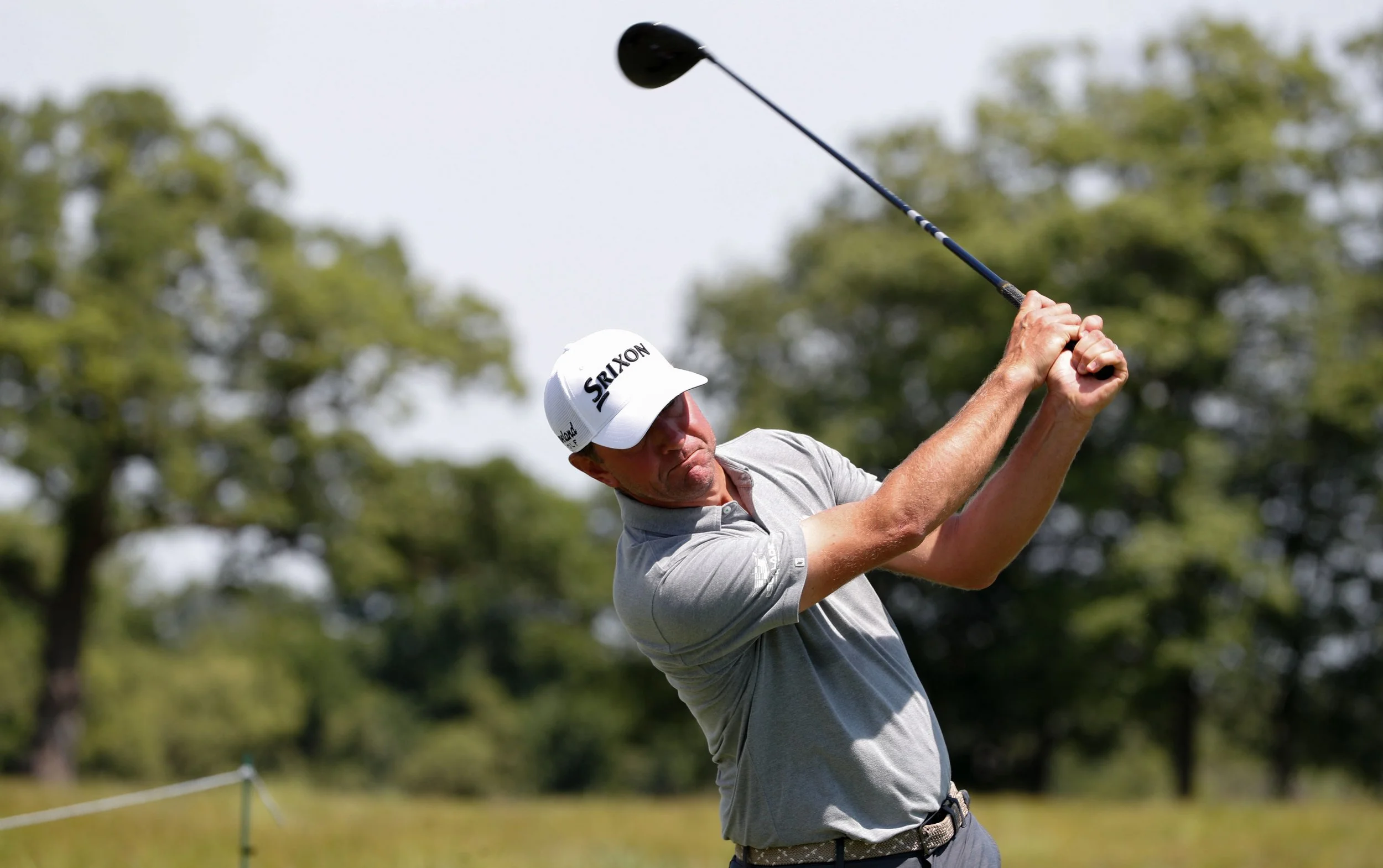 A man wearing a gray polo shirt, white cap, and sunglasses is swinging a golf club on a golf course with green trees in the background.