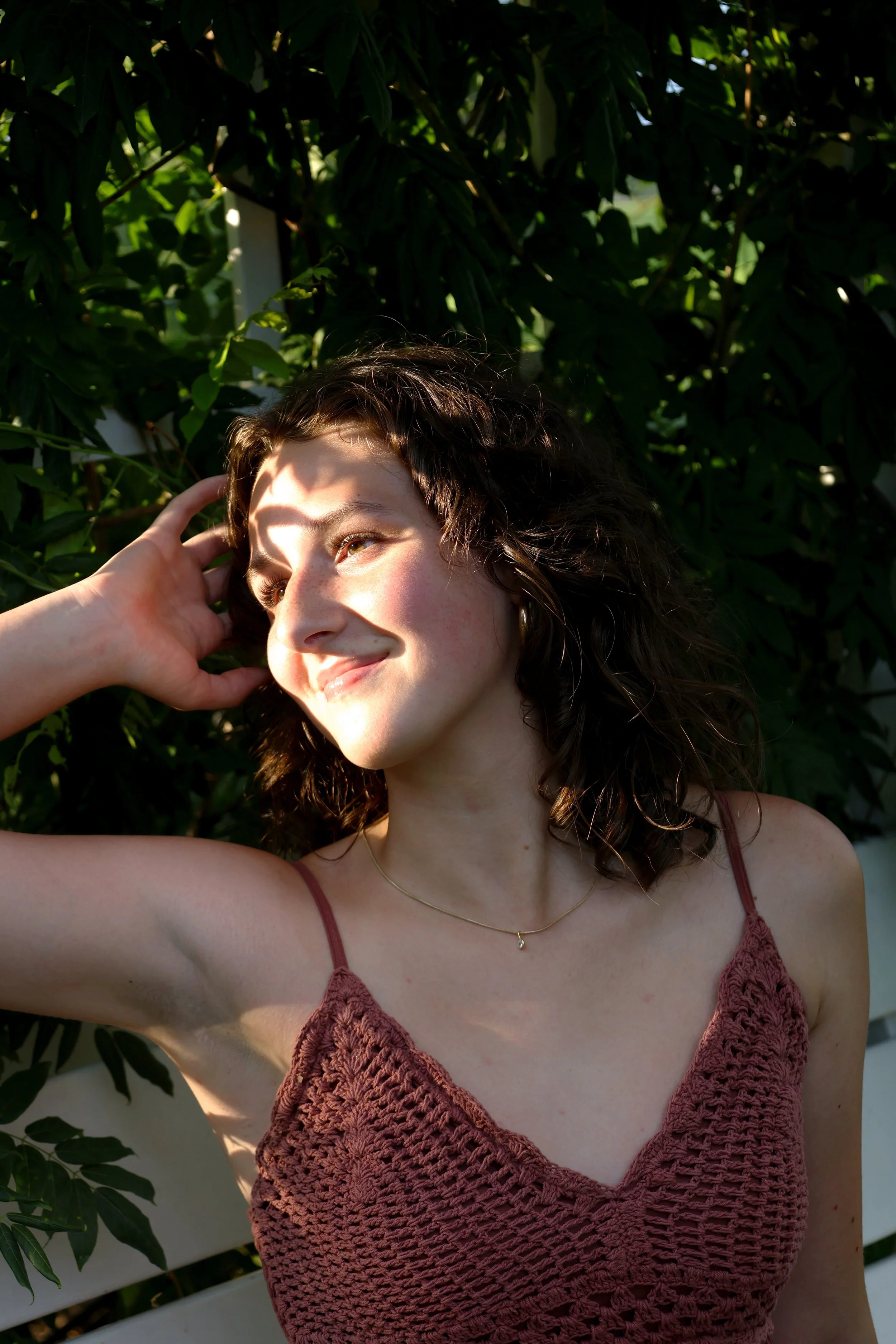 A young woman with curly brown hair smiling and looking slightly upward, wearing a maroon crochet top and a delicate gold necklace, standing in front of green leafy plants with sunlight creating shadows on her face.