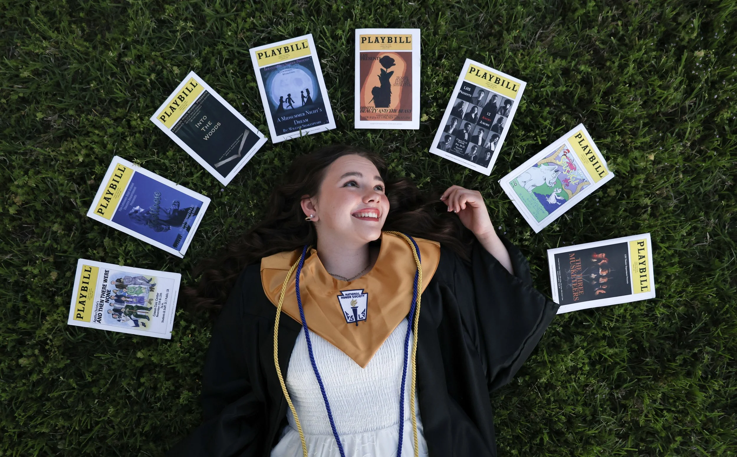 A young woman lying on green grass, dressed in a graduation gown and cap, smiling with her eyes open, surrounded by multiple playbills for various theater productions.