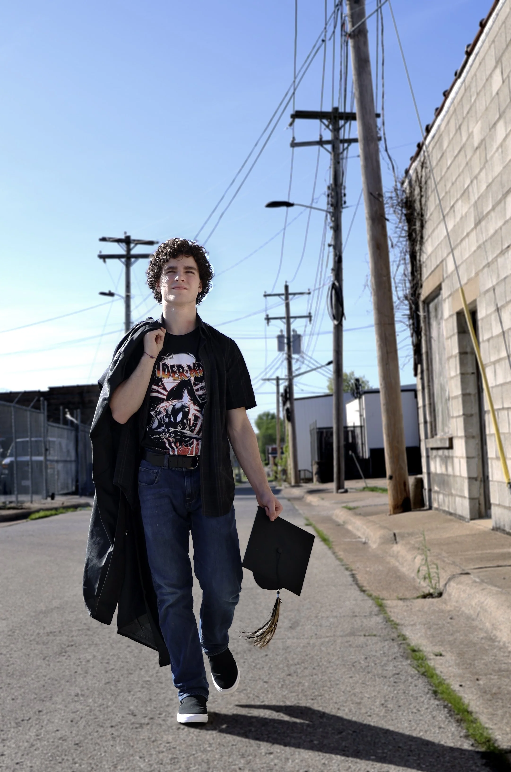 A young man with curly hair walking outside with a graduation cap in his right hand and a black gown over his shoulder, wearing a black T-shirt featuring graphic art, jeans, and sneakers, on a sunny day near utility poles and a brick building.
